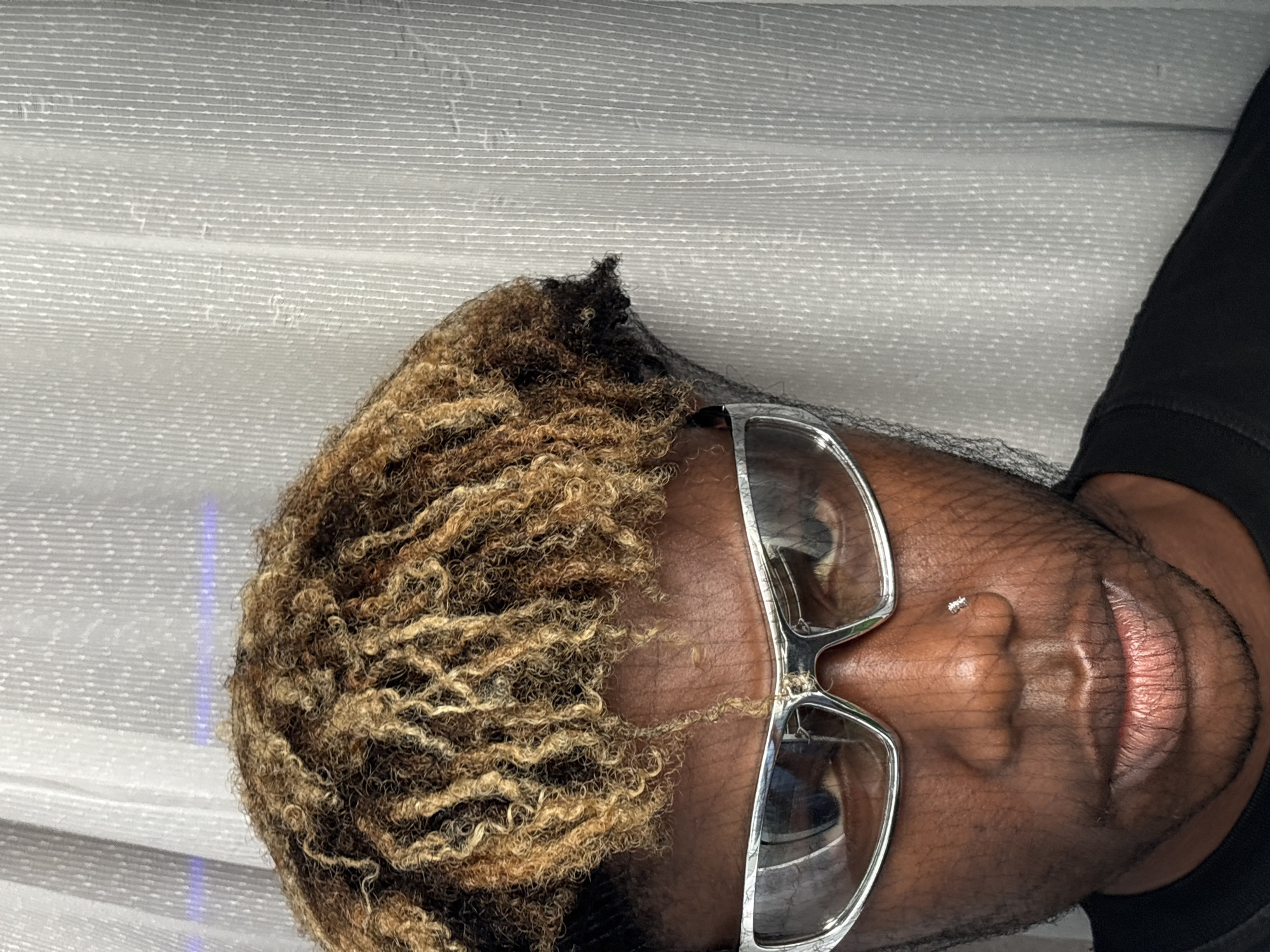 The image displays a close-up portrait of a person, likely male, with a dark skin tone, captured from the chest up. The photograph is oriented 90 degrees counter-clockwise. The subject has light brown and blonde curly hair, with visible darker roots, and their hair is covered by a black net. They are wearing eyeglasses featuring thick, silver-colored frames and reflective lenses. A small, shiny piercing is visible on the left side of their nose, and their full lips are slightly parted. A black garment, possibly a shirt or top, is visible on the right side of the image.
The background is a light-colored, textured surface characterized by subtle horizontal lines, suggesting an indoor setting, possibly fabric or blinds. A faint, vertical streak of blue light is present on the left side of the background. The person appears to be posing for the camera. There is no visible text in the image. The lighting is soft and even, highlighting the subject's features. The location context for this image is Igboefon, Nigeria.