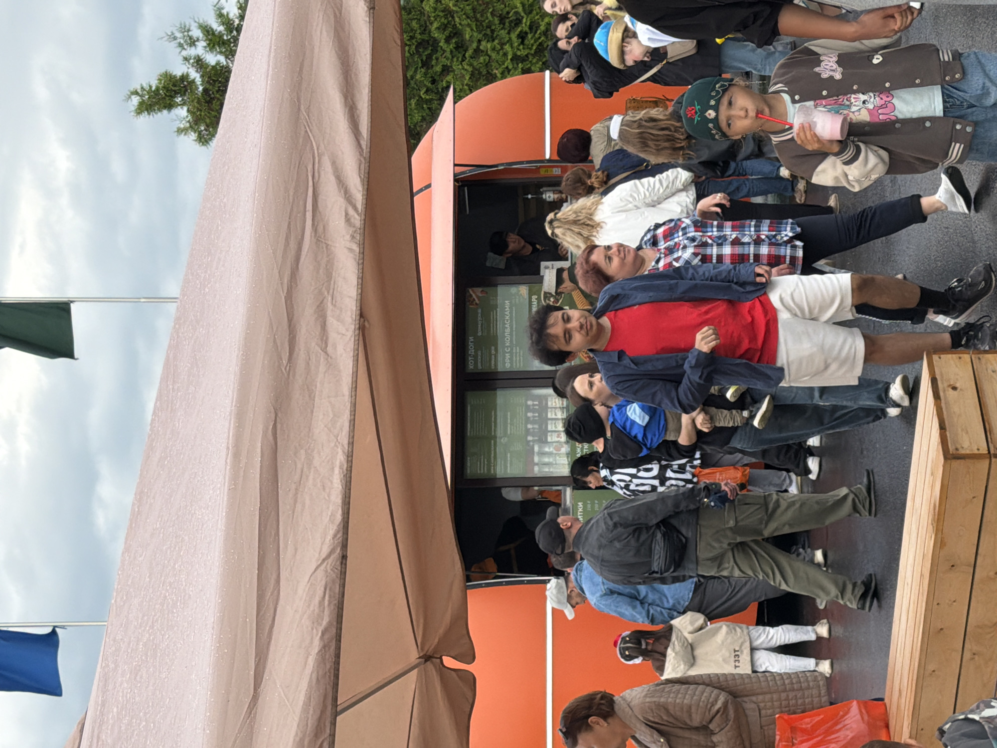 The image depicts a crowd of people standing in line, seemingly outside a food truck or a stand with a menu. The scene appears to be outdoors, under an overcast sky, and there are some signs of recent rain, indicated by water droplets on the tent fabric. The overall mood seems relaxed, with people patiently waiting.
Several individuals are visible, dressed in casual attire. Some are looking towards the food stand, while others are engaged in conversation or looking at their phones. A young person in the foreground is holding a pink drink with a straw.
The food stand has an orange exterior and a large, open window where orders are likely being taken. A menu board is visible inside the window, with some text in Cyrillic script. Some legible text from the menu includes "хот-доги" (hot dogs), "французема" (French fries), and "ФРИ С КОЛБАСКАМИ" (fries with sausages). The price "250 P" is also visible.
A large beige tent covers part of the scene, suggesting it might be a temporary setup for an event or a business. Two flags are also visible, one green and one blue, attached to poles in the background. The presence of a wooden bench suggests a waiting area. The time of day appears to be daytime, given the ambient light, but the cloudy sky makes it difficult to be precise. The overall impression is that of a casual outdoor food service or gathering.
