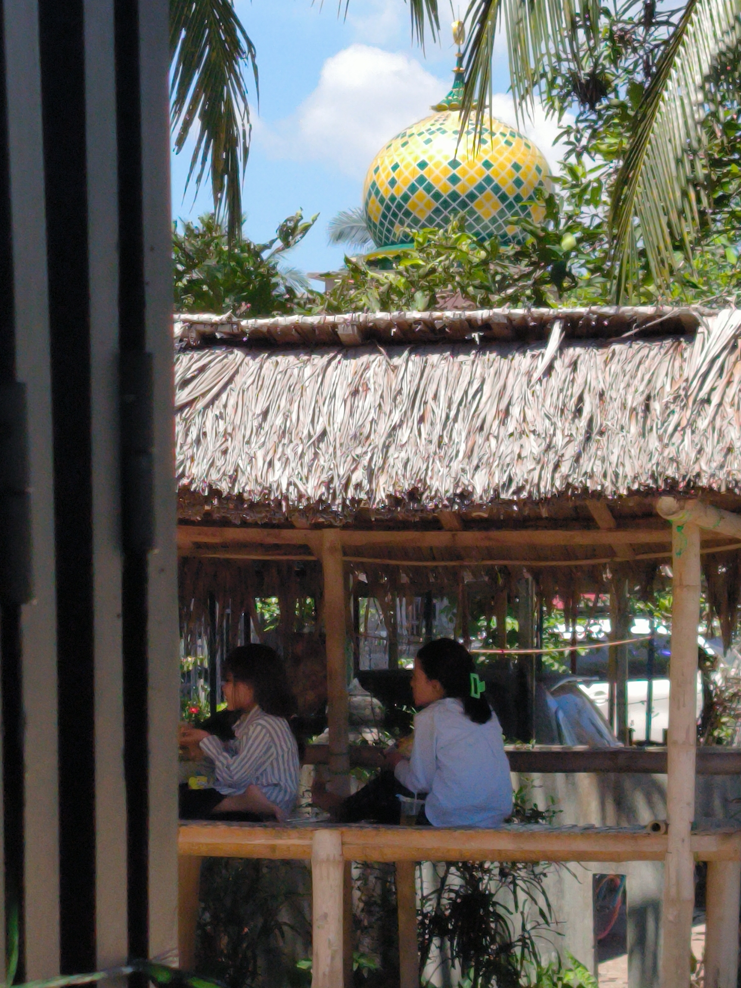 Two children are seated on a raised platform structure with a thatched roof in Merbopuntong, Indonesia. A yellow and green patterned dome is visible above the roofline, suggesting a nearby religious building. The structure is constructed of light-colored bamboo or wood. The children appear to be engaged in an activity that involves their hands. A partially obscured fenced structure is located in the foreground, and foliage surrounds the structure. The sky is partially visible in the background.
