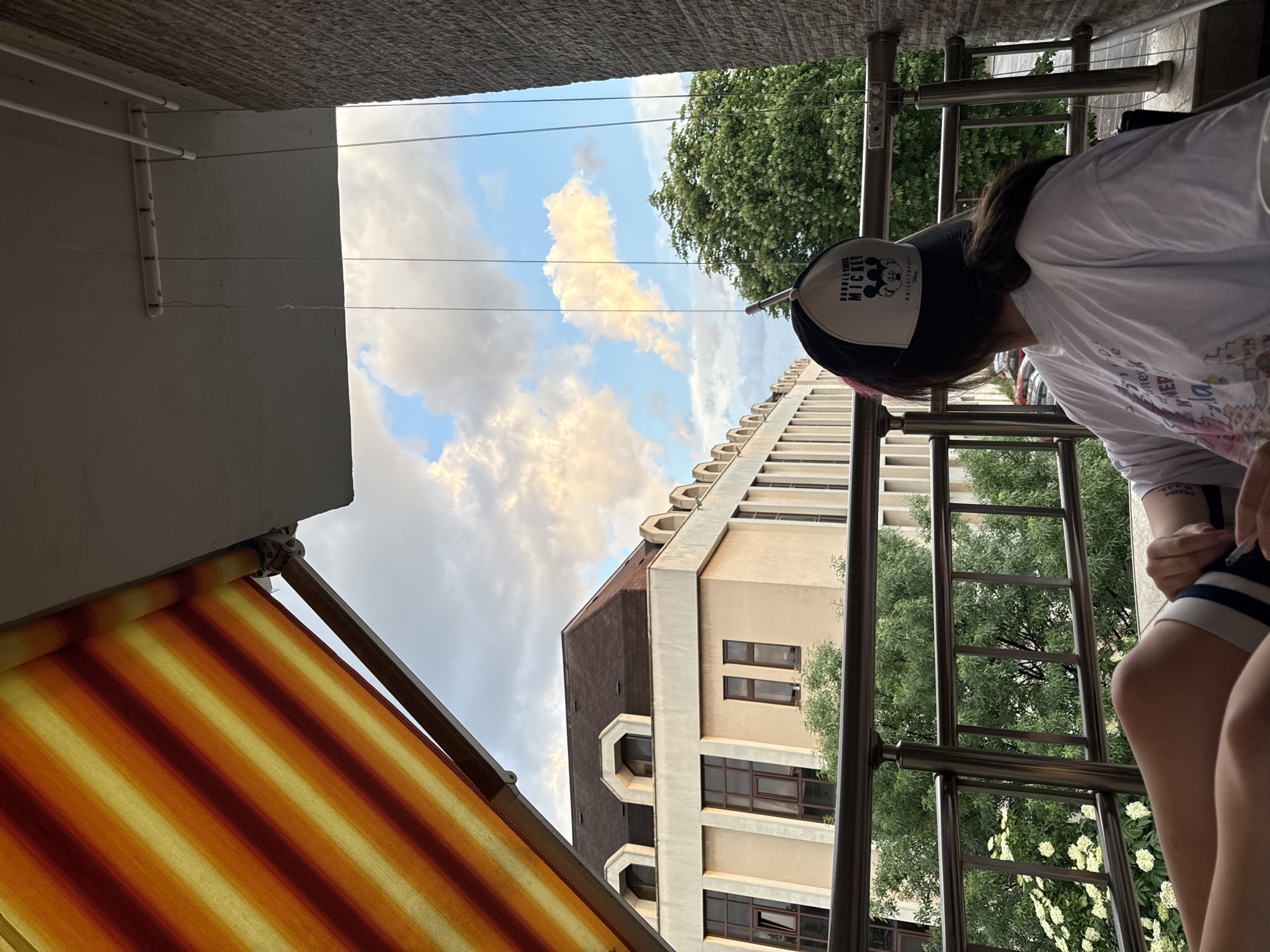 A low-angle shot taken from what appears to be a balcony in Sofia, Bulgaria. A person wearing a black and white "Double Scoop Mickey" baseball cap and a white t-shirt stands on the right. The person is near a metal railing overlooking the street. In the background, a tall beige building with a brown roof is visible, framed by green trees and a sky dotted with clouds. On the left, a striped orange and yellow awning partially obstructs the view. Overhead, a gray concrete ceiling is visible, with clotheslines.