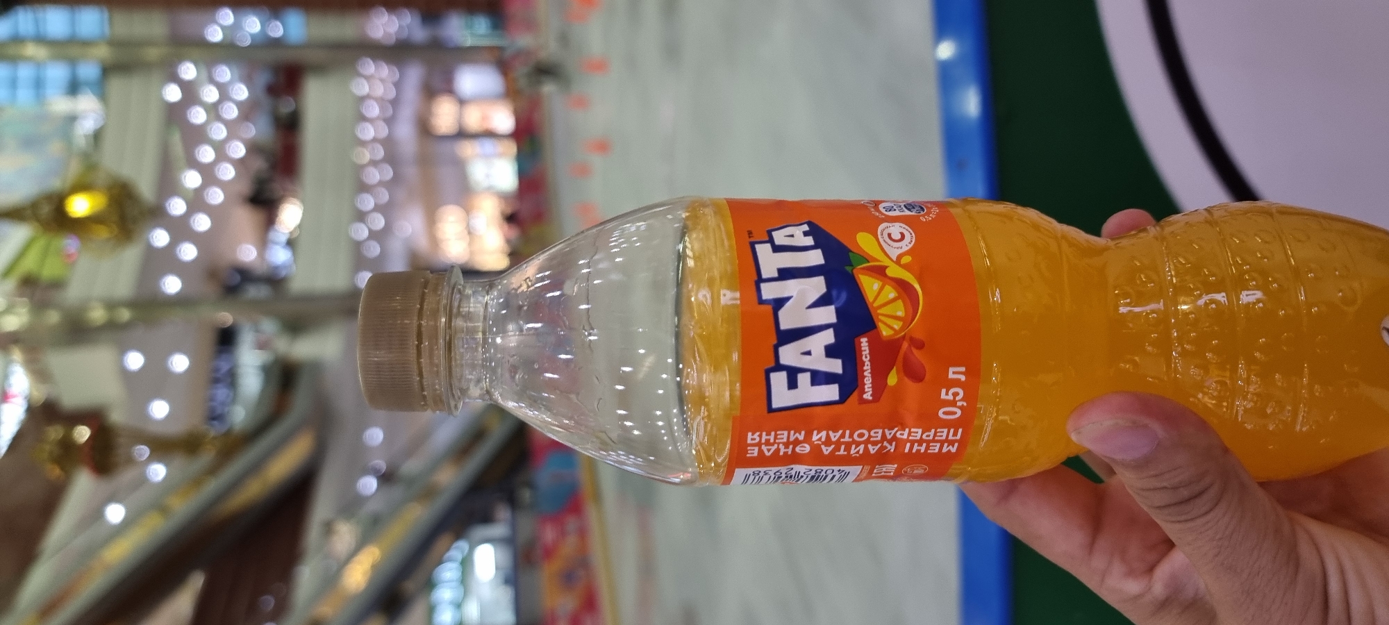 A close-up, eye-level shot shows a hand holding a 0.5-liter bottle of Fanta orange soda. The bottle is clear plastic and filled with the orange beverage. The label on the bottle is primarily orange with the Fanta logo in blue and white. The background is out of focus, showing a brightly lit indoor space with escalators and decorative lights, suggesting a shopping mall or entertainment venue. The lighting appears to be a mix of artificial and natural light, creating a warm glow. The overall impression is one of casual enjoyment in a public setting. This scene could be situated in Aktobe, Kazakhstan, a city known for its modern shopping centers and recreational facilities.