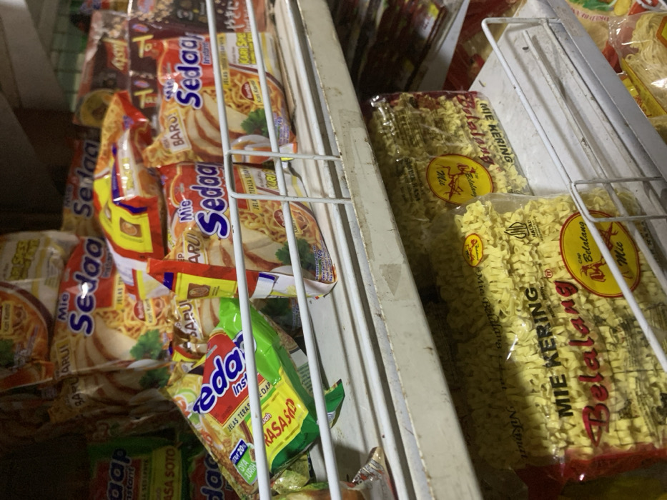The image shows the interior of a refrigerator, stocked with various packaged instant noodles. The refrigerator is white with rusty metal shelves. On the left side, there are multiple packages of "Mie Sedap" brand instant noodles with different flavors, all featuring a red and yellow color scheme and images of noodles, chicken, and vegetables. On the right side, there are two packages of "Mie Belalang" brand instant noodles with a yellow and red color scheme and images of noodles and a logo. The noodles in the packages appear to be dry, curly, and yellow. The packages are arranged vertically and horizontally on the shelves, with some partially obscured by others. The image was taken in a store in Kecamatan Lawang, Indonesia, located at latitude -7.829 and longitude 112.696.