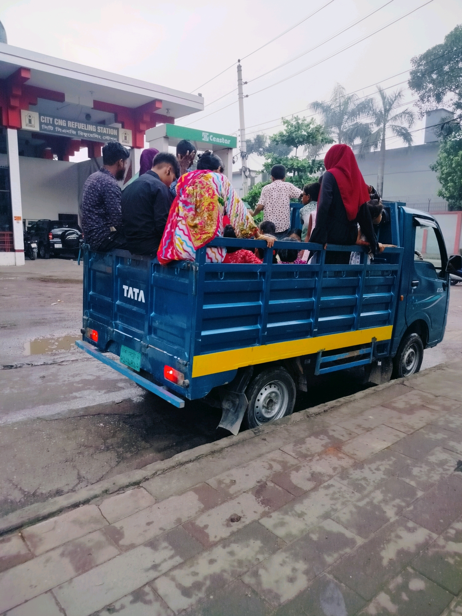 In Dhaka, Bangladesh, a blue Tata pickup truck, carrying several passengers, is partially submerged in a ditch or pothole on a wet, paved road. The truck's right-side wheels are deep in the ditch, and the vehicle appears to be stuck. On the truck bed, multiple individuals, including men, women, and children, are seated or standing. Some are wearing traditional clothing, and one woman is prominently visible in a red hijab. The truck has "TATA" branding on its tailgate. In the background, a "City CNG Refueling Station" sign is visible, suggesting the location is near a gas station. The sky is overcast, and the surrounding environment appears to be an urban setting with some greenery and buildings. The road surface is wet and shows signs of wear and tear, with visible puddles and cracks.