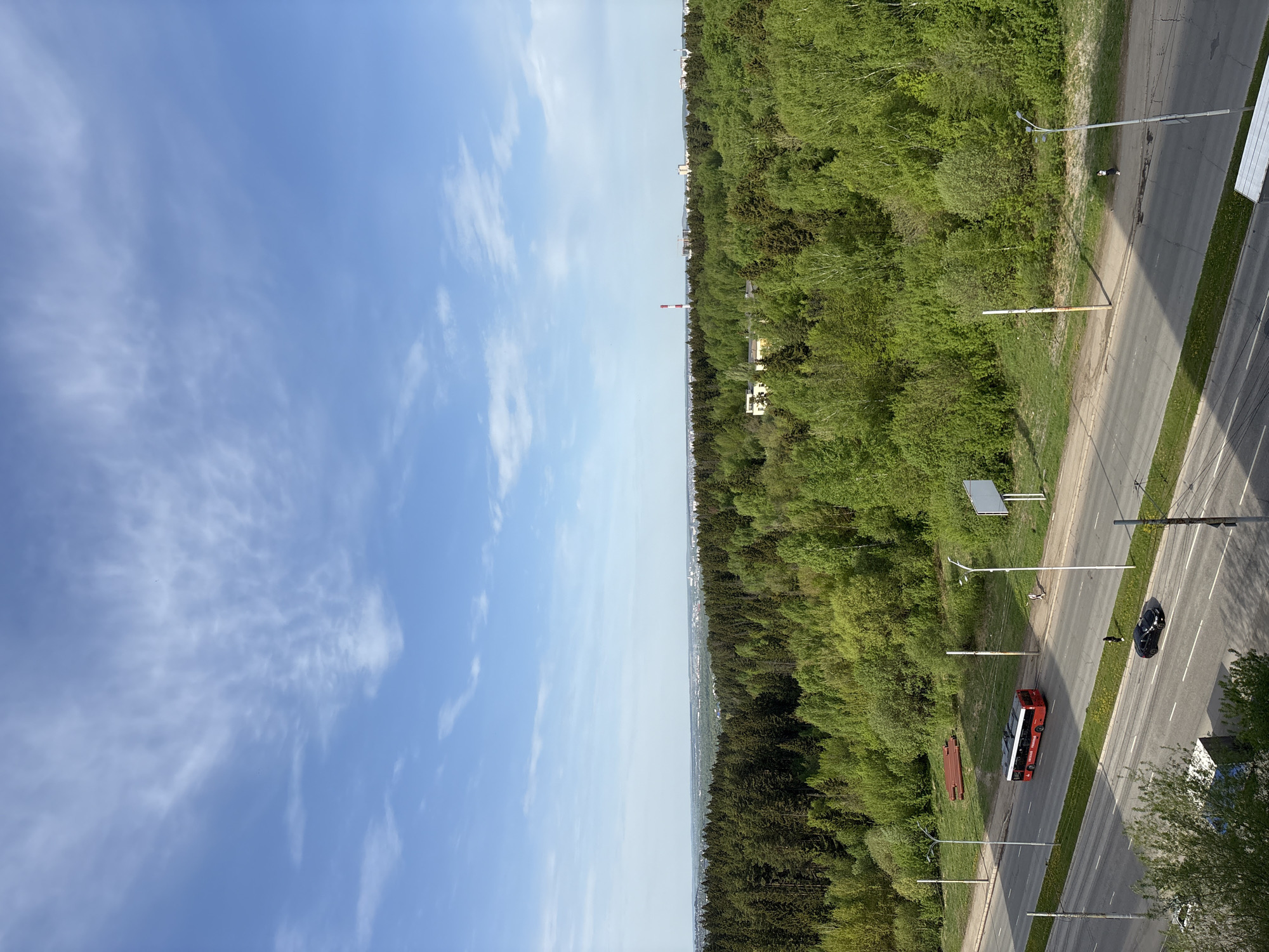 The image depicts an elevated view of a scene in Izhevsk, Russia. The left half of the image is predominantly a blue sky with scattered clouds. The right half shows a dense green forest transitioning to a road. On the road, a red bus is parked, and a dark-colored car is moving away. The road is flanked by sidewalks, lampposts, and some greenery. In the distance, buildings can be seen above the forest line, and a tall chimney is visible.