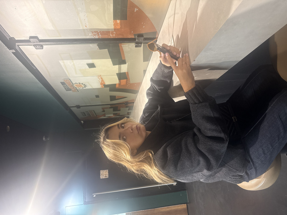 An interior image depicts a female individual seated at a counter, likely within the lobby or snack area of a UGC Ciné Cité cinema, specifically UGC Ciné Cité Carré de Soie in Vaulx-en-Velin, France, as indicated by the "UGC FLAMMES" logo on the background wall. The subject, a woman with shoulder-length blonde hair, appears to be in her 20s or 30s and is wearing a dark gray, high-collared jacket or sweater, dark blue jeans, and a dark chain-strap shoulder bag. She is positioned on a round, brown stool, facing the camera with a neutral expression. Her left hand holds a dark smartphone, while her right hand holds a pair of brown-framed sunglasses, both resting on a light gray, possibly concrete, counter. The background features a wall composed of large panels adorned with a muted orange, beige, and black geometric graphic design. To the left, a dark door with a white restroom symbol is visible. The ceiling is dark, with recessed lighting, and a prominent lens flare is present in the upper left quadrant of the image.