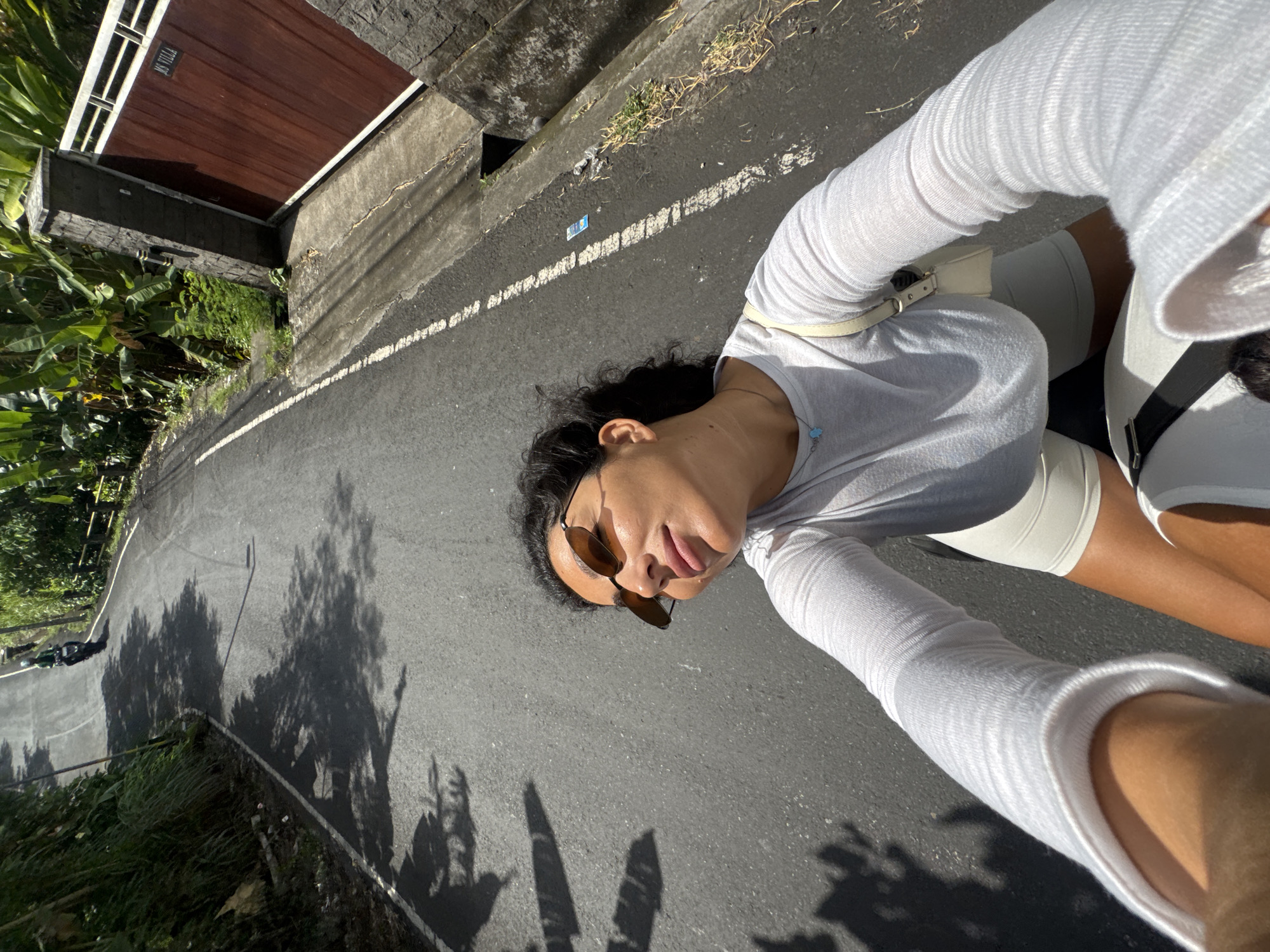 A woman wearing sunglasses and a white long-sleeved shirt is riding a scooter down a paved road lined with lush greenery and trees. Her head is tilted back, and she is looking upwards. The road has a white dashed line marking the lane. In the background, a brown wooden gate with "MS VILLA" written on it is visible, along with a stone pillar. Sunlight casts shadows of trees on the road. A person on another scooter is further up the road. The scene appears to be in a tropical environment, consistent with locations in North Kuta, Indonesia.