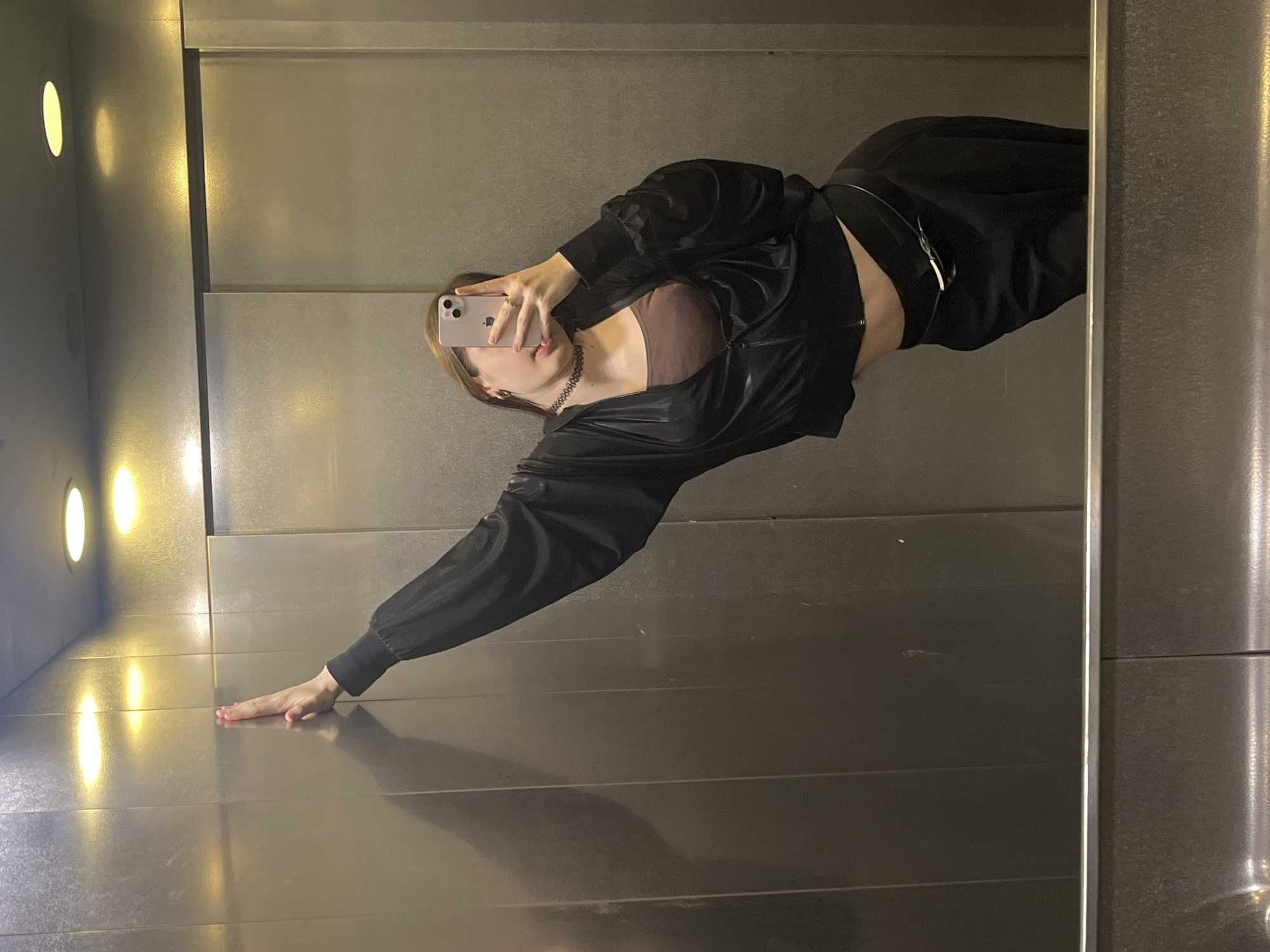 The image shows a woman taking a selfie in what appears to be an elevator. She is dressed in black clothing, including a jacket, and holds a smartphone in her right hand, obscuring part of her face. Her left hand is extended and resting on the elevator floor. The elevator's interior consists of metallic walls and integrated lighting. The location is not explicitly discernible from the image. There are no apparent objects, actions, or interactions beyond the described pose. There is no evidence to suggest that the scene is in Saint Petersburg, Russia.