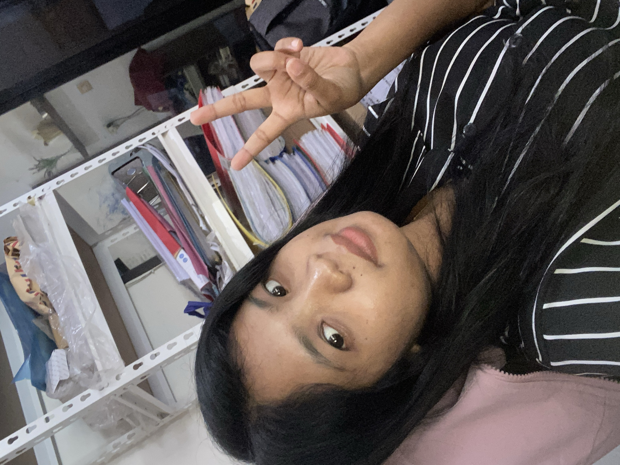 A person with dark hair is shown from the chest up, wearing a black and white striped shirt over a light pink garment. They are making a "peace" or "victory" sign with their right hand. Behind the person is a white metal shelving unit. The shelving unit is partially filled with file folders and objects wrapped in plastic. A dark reflective surface, possibly glass or polished stone, is visible above the shelving unit. It is unknown if this picture was taken in Negara, Indonesia.