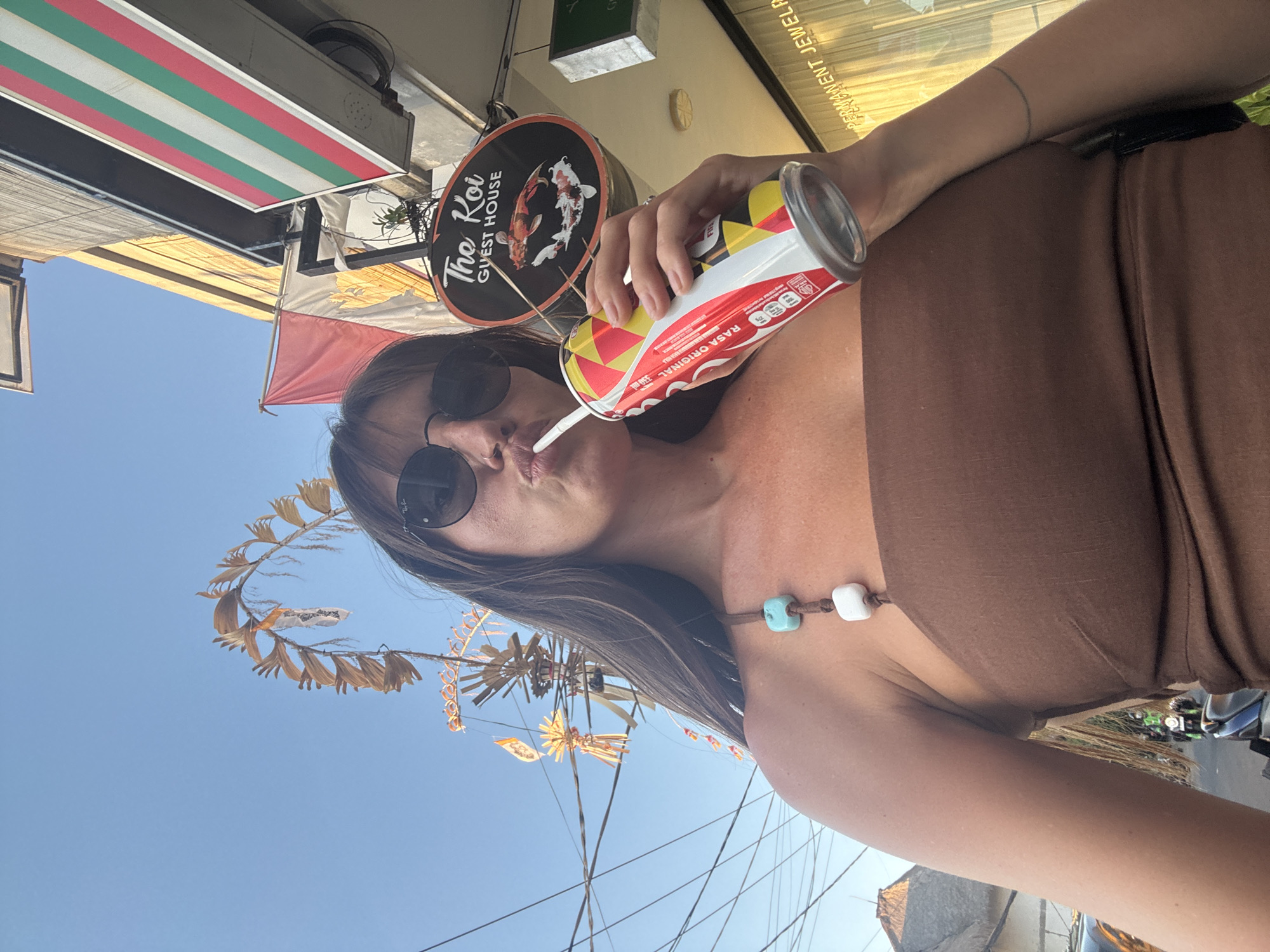 In North Kuta, Indonesia, a woman with long dark hair and sunglasses is captured in a selfie, holding a can of Coca-Cola. She is wearing a brown tube top with a beaded strap featuring turquoise and white beads. The background reveals a street scene with buildings, including a sign for "The Koi Guest House" with two koi fish illustrations. A red and white Indonesian flag is visible on a pole, along with other decorative elements and overhead wires. The woman's lips are pursed as she sips from the can through a straw. Further down the street, a motorcycle is seen in motion, suggesting a lively urban environment. The image is taken outdoors during the daytime, with a clear blue sky.