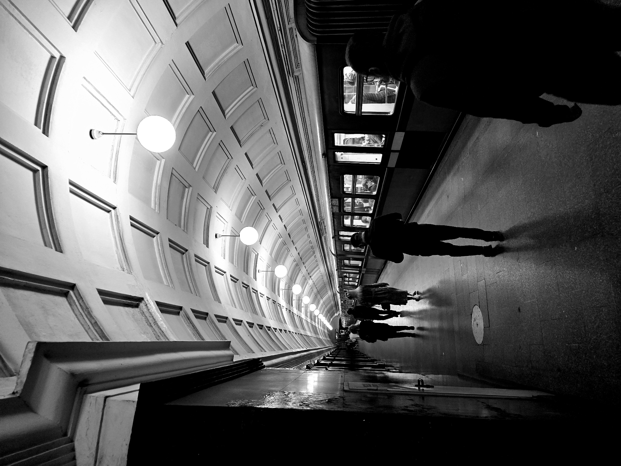 The black and white image depicts a subway station platform in Moscow, Russia. The architecture features a curved, tiled wall with evenly spaced globe-shaped light fixtures extending into the distance. The platform is occupied by several people, mostly silhouettes, appearing to be waiting for or boarding a train. Some are standing, and others appear to be in the train. The reflective, tiled floor amplifies the perspective and creates a sense of depth. A train is visible along the right side of the frame.