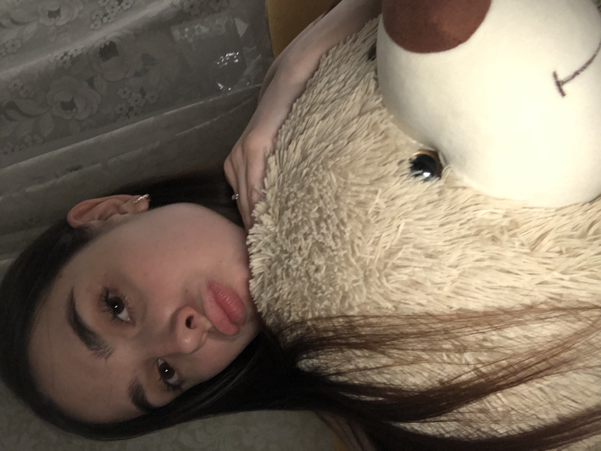 A young woman with dark hair lies on a surface with a lace pattern. She holds a large, fluffy stuffed animal, predominantly beige with a white head and a brown patch. The woman is wearing a ring on her left hand and has an earring in her left ear. She is making a pouting expression. No information exists that confirms that this image was taken in Noviy, Russia.