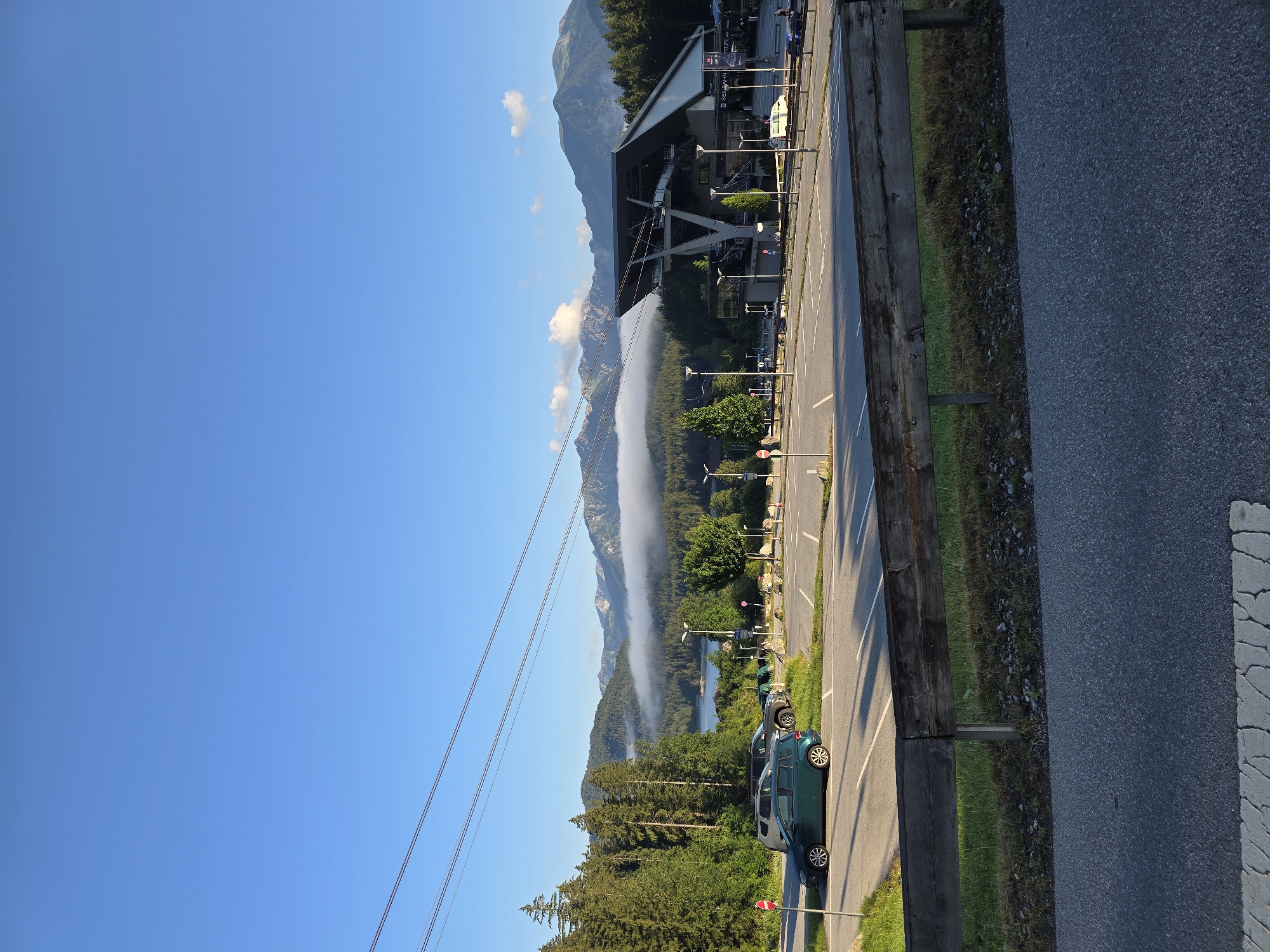 The image depicts a scenic view in Grainau, Germany, featuring a cable car station in the foreground against a backdrop of mountains and a lake. The cable car station, a modern structure with a prominent roof, is situated on the right side of the frame. In the foreground, a paved parking lot is visible with several cars parked. A wooden barrier runs across the bottom of the parking lot, separating it from a road in the very front. The middle ground is dominated by a lush green forest, with tall pine trees lining the slopes. Beyond the forest, a tranquil lake is nestled, reflecting the clear blue sky. The background is defined by a range of majestic mountains, their peaks partially shrouded in mist, adding a dramatic effect to the landscape. The sky is a brilliant, cloudless blue, indicating fair weather. The overall scene is one of natural beauty and outdoor recreation, typical of a mountain resort town.