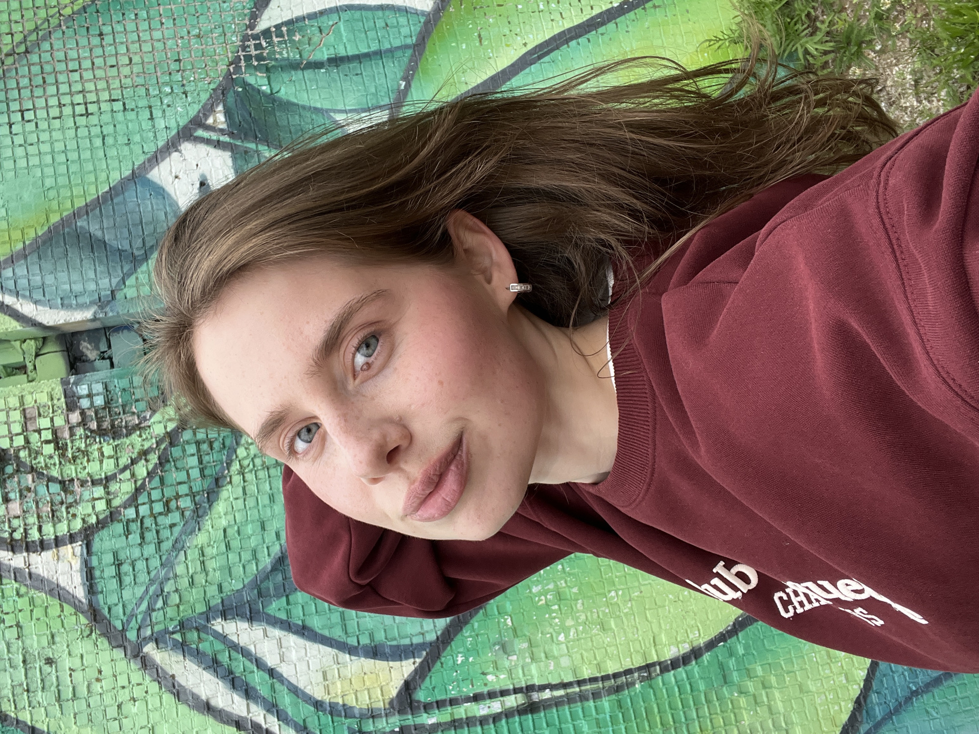 The image shows a young woman lying down and taking a selfie against a green and white graffiti-covered wall. She has long, brown hair that is spread out, and she is wearing a dark red sweatshirt. Her face is turned towards the camera, and she has a slight smile. She is wearing a small, rectangular earring in her left ear. The background features vibrant green graffiti with some white abstract shapes and a grid-like texture. A patch of grass is visible in the upper right corner. The lighting suggests it might be daytime, possibly outdoors. The setting appears to be urban or a place with public art. There is text visible on her sweatshirt, which appears to say "hub" and "Chaue" in white capital letters. The woman's expression is relaxed and pleasant. The overall mood of the image is casual and contemporary.