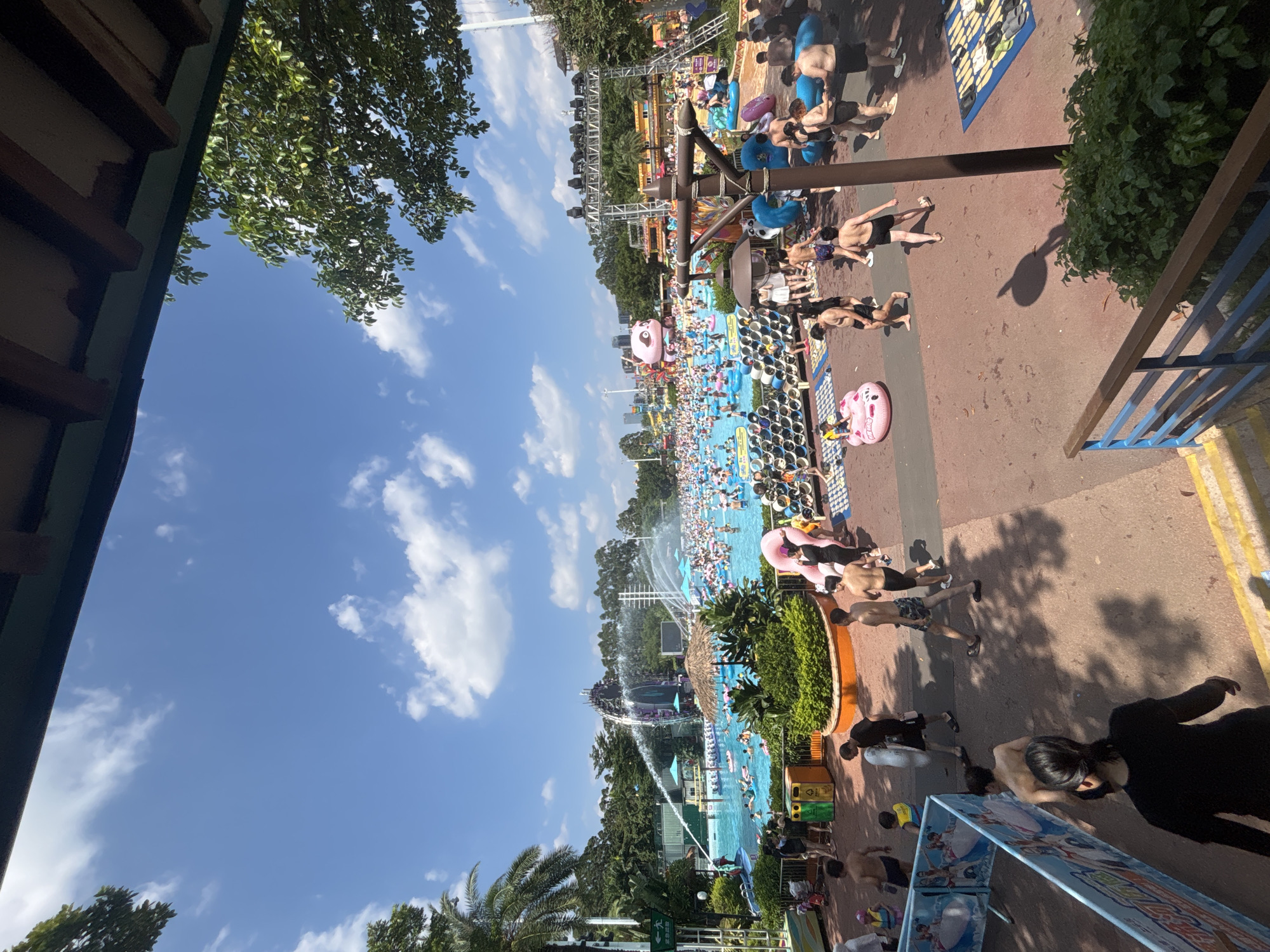 The image depicts a crowded water park on a sunny day. A large pool is filled with people of all ages, many of whom are floating on inflatable rings or tubes. Water jets are spraying from a central structure, creating a lively atmosphere. In the background, a large inflatable pink panda figure is visible, along with various water slides and attractions. Palm trees and lush green foliage surround the pool area, suggesting a tropical or sub-tropical climate. People are seen walking around the perimeter of the pool, some in swimwear, while others are relaxing on lounge chairs or seated in shaded areas. The overall scene conveys a sense of fun and recreation. The provided hint images confirm that this water park is located in Panyu District, China.