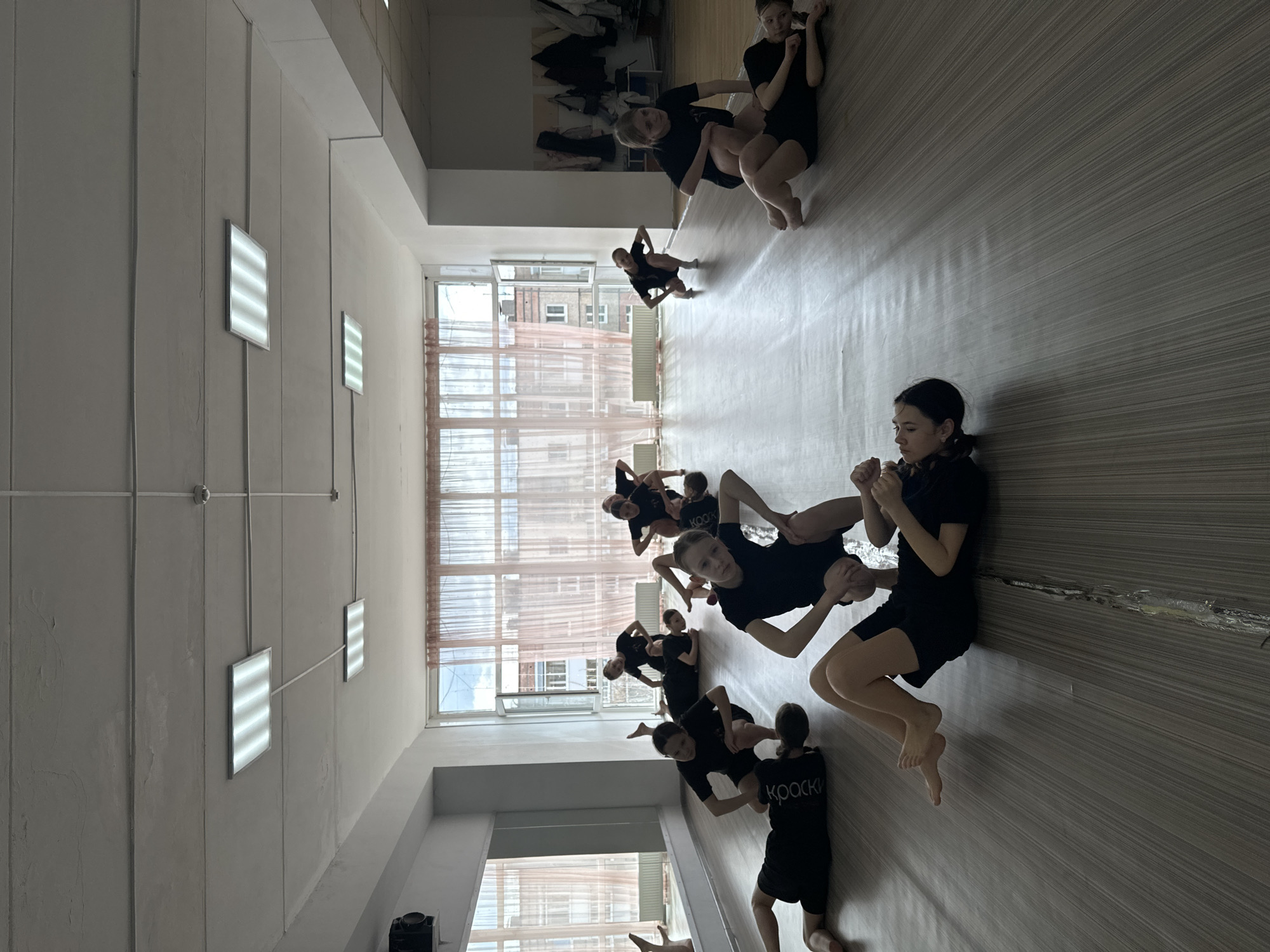 The image displays an indoor scene, likely a dance or gymnastics studio, where a group of young girls are participating in a physical training session. Approximately ten to twelve girls, appearing to be school-aged, are visible. They are uniformly dressed in black short-sleeved tops and shorts or leotards. The girls are spread across a light-toned floor, which features a subtle striped pattern, engaged in various stretches, poses, and partner exercises. Some are seated, leaning on each other, or holding hands, suggesting cooperative drills.
The room has high ceilings equipped with several rectangular fluorescent light fixtures that provide artificial illumination. Large windows line one side of the space, allowing ample natural light to fill the room. Through these windows, residential or urban buildings are visible outside, partially obscured by sheer, light-colored curtains. The bright light indicates it is daytime.
Notable text is visible on the backs of some of the girls' shirts. One shirt clearly displays the Cyrillic word "КРАСКИ" (KRASKI), which translates to "PAINTS" or "COLORS." Another shirt features the text "KOOKKO." A black speaker or audio device is situated on the floor near the windows. The overall atmosphere suggests focused activity, with the girls appearing concentrated and engaged in their practice within this functional and well-lit training facility.