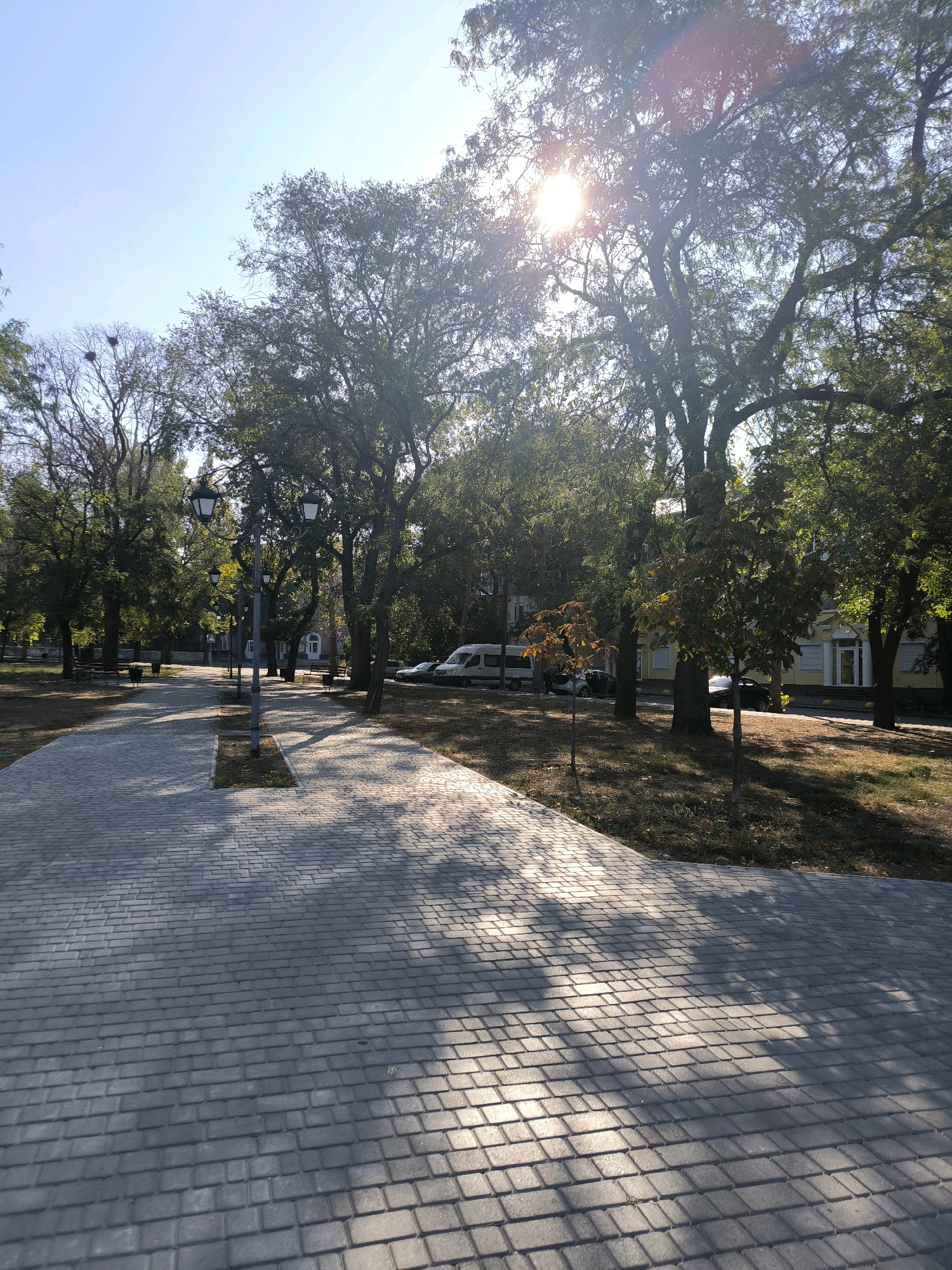 This is an outdoor, eye-level shot of a paved walkway in a park on a sunny day. The walkway is made of grey interlocking pavers and is lined with trees on both sides. The sun is shining brightly through the trees, casting dappled shadows on the walkway. In the background, there are buildings and parked vehicles, including a white van and a dark sedan. The scene appears to be in Mykolaiv, Ukraine, based on the context provided. There are no people visible in the image.