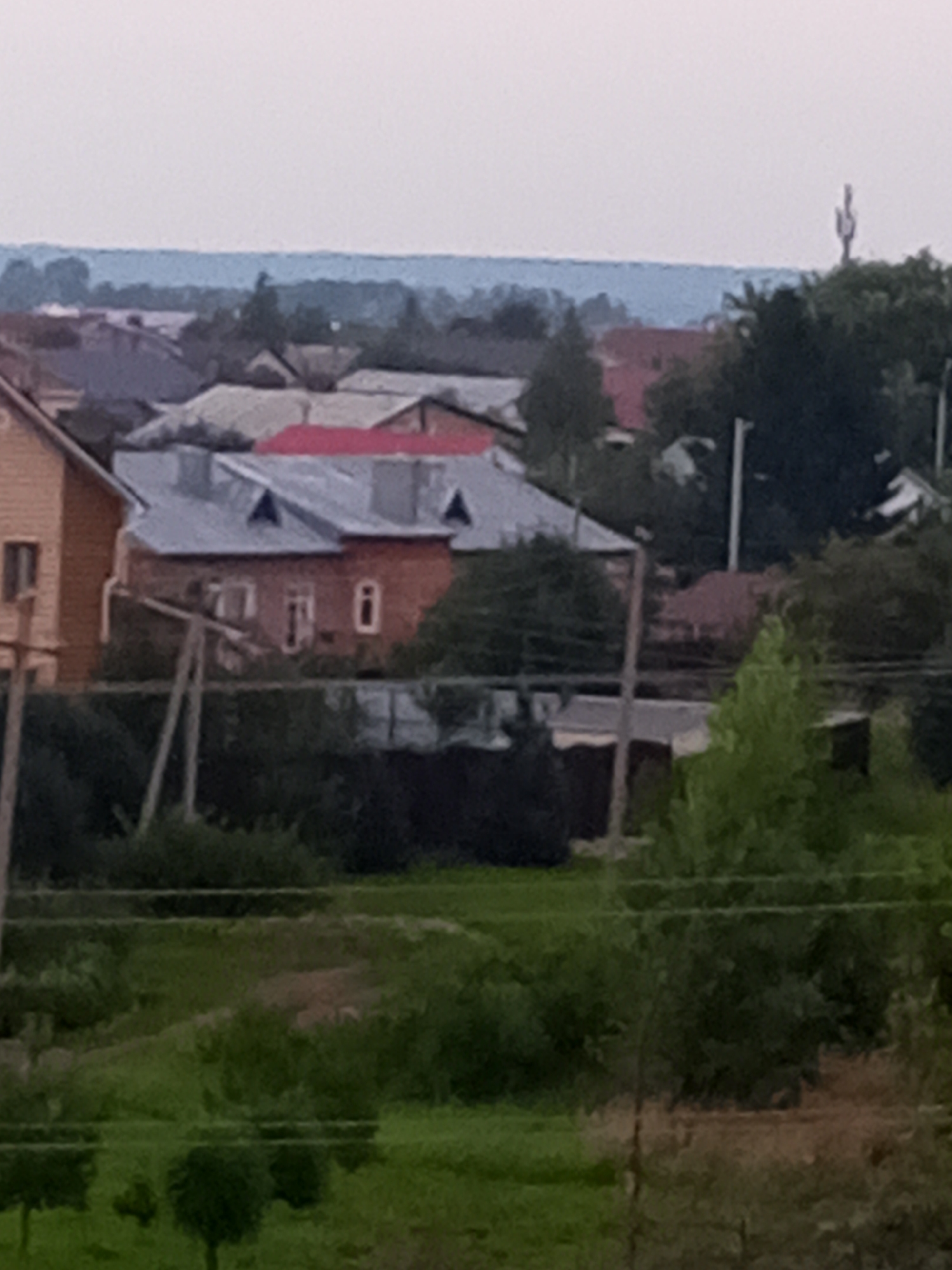 The image is a low-resolution, slightly blurry photograph of a residential area, likely in a rural or suburban setting. In the midground, a two-story building with a red roof and multiple windows is visible. Adjacent to it is another building with a gray roof and a single window. The foreground shows lush green vegetation, including trees and bushes. In the background, a hazy blue horizon suggests distant hills or a large body of water. The sky is overcast. The overall impression is one of a quiet, possibly overcast day in a village or small town. The specific location of Chistopol, Russia, is not discernible from the visual information in the image itself.