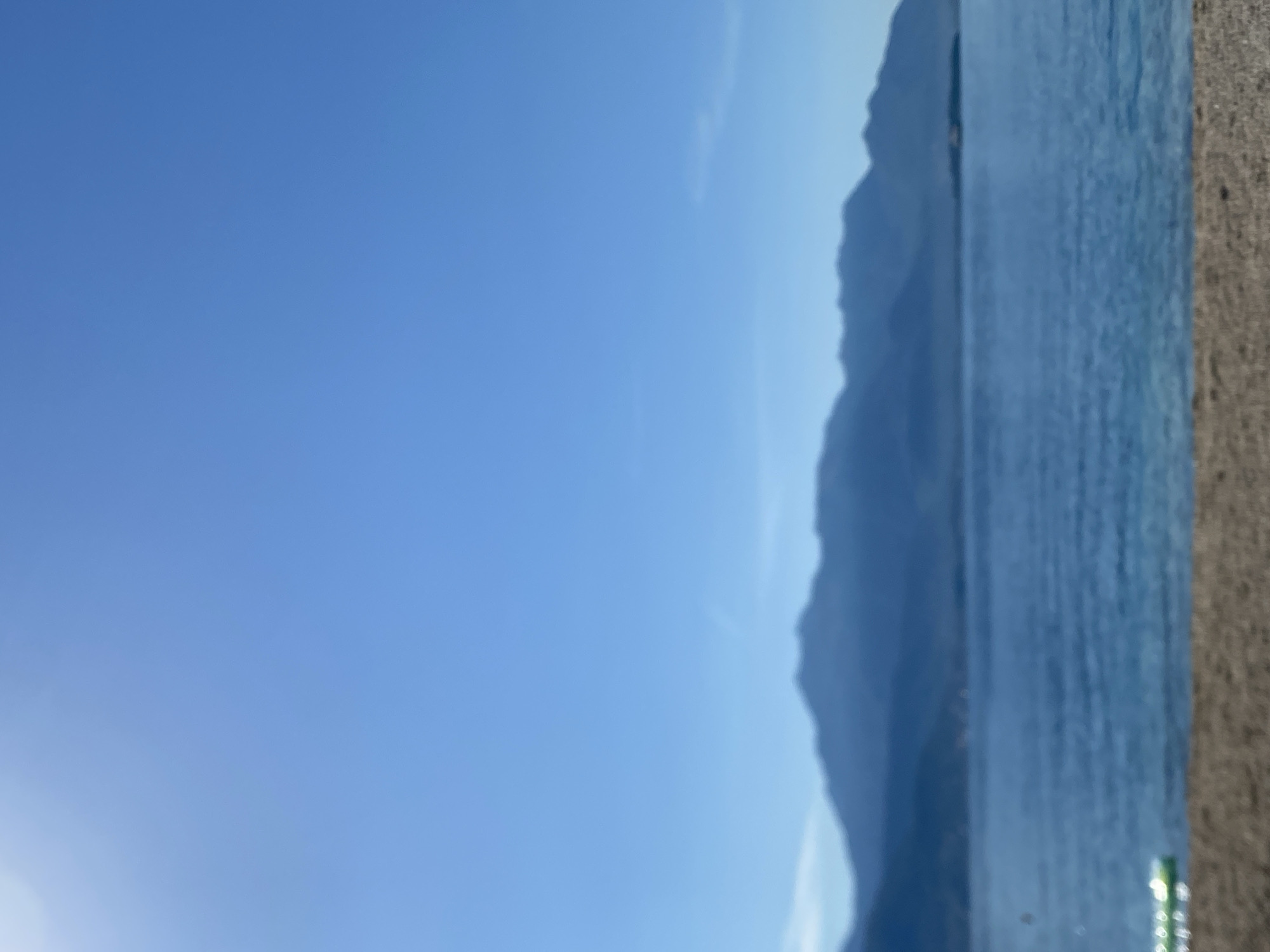 The image depicts a serene coastal scene. In the foreground, a sandy beach is visible. Beyond the beach lies a calm body of blue water, likely the Adriatic Sea. In the distance, a range of hazy, dark blue mountains forms the horizon. The sky above is a clear, bright blue with a subtle sun glare in the upper left corner. A small, partially submerged inflatable object with green and white markings is partially visible in the lower left corner of the water. The scene is tranquil and appears to be set in the city of Baošići, Montenegro, a coastal town known for its beautiful natural scenery.