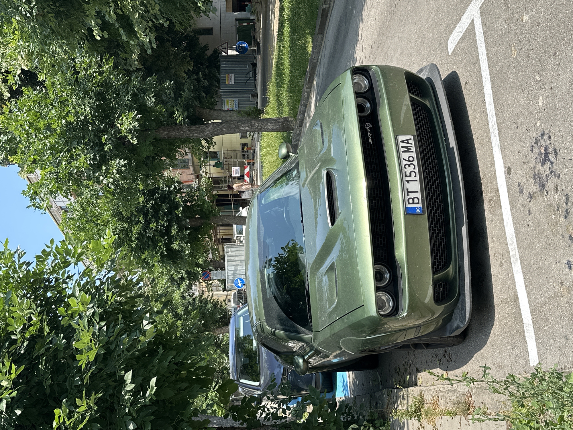 The media file captures a front-end view of a green Dodge Challenger SRT parked on an asphalt surface, likely a street or parking lot, in Veliko Tarnovo, Bulgaria. The car is angled towards the viewer, with its distinctive grille, headlights, and hood scoop prominently displayed. A license plate with the text "BT 1536 MA" and a blue and yellow BG symbol is visible. The car is parked within white painted lines, suggesting it is in a designated parking spot.
The setting is outdoors on a sunny day, indicated by the bright lighting and strong shadows. Lush green trees and foliage border the scene, with some leaves casting dappled shadows on the car and the ground. In the background, there are glimpses of buildings and what appears to be a construction site with scaffolding and signs. The overall atmosphere is that of a typical urban or suburban environment on a clear day. No people are clearly visible, though the environment suggests pedestrian activity. The image focuses primarily on the vehicle, highlighting its design and presence within its surroundings.