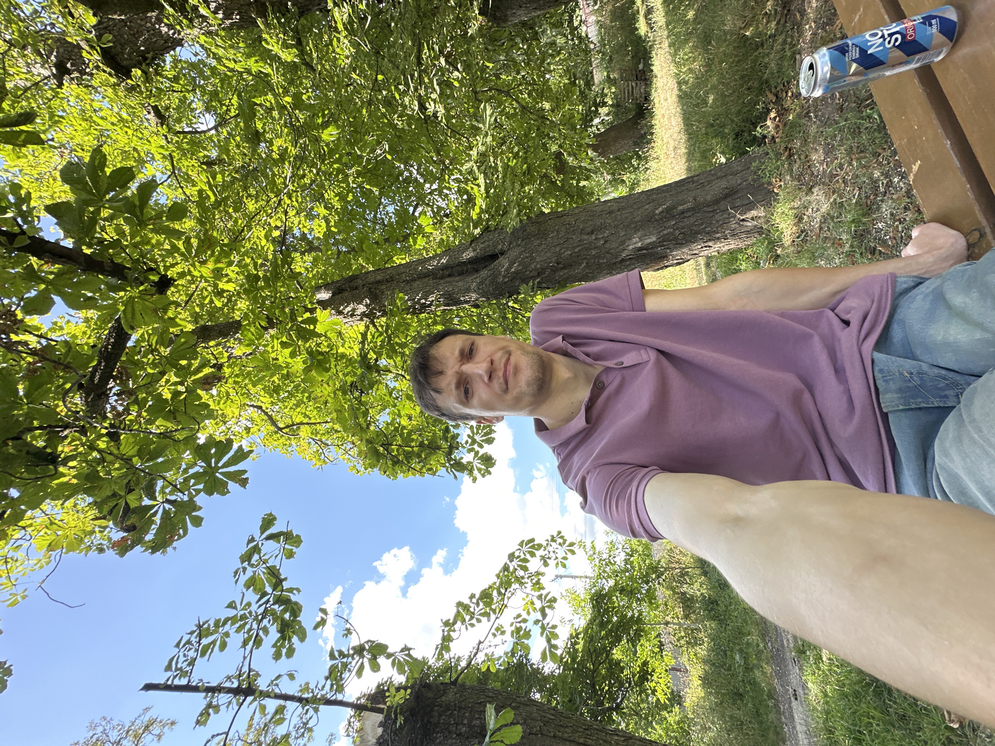 A young Caucasian man is sitting on a wooden bench in a park. He is wearing a purple polo shirt and blue jeans. His right arm is extended towards the camera, as if taking a selfie. To his right, there is an open can of energy drink. The background is filled with lush green trees and a bright blue sky with white clouds. The man's facial expression is neutral, and he is looking directly at the camera. The park appears to be in the city of Makiv, Ukraine, based on the provided information.