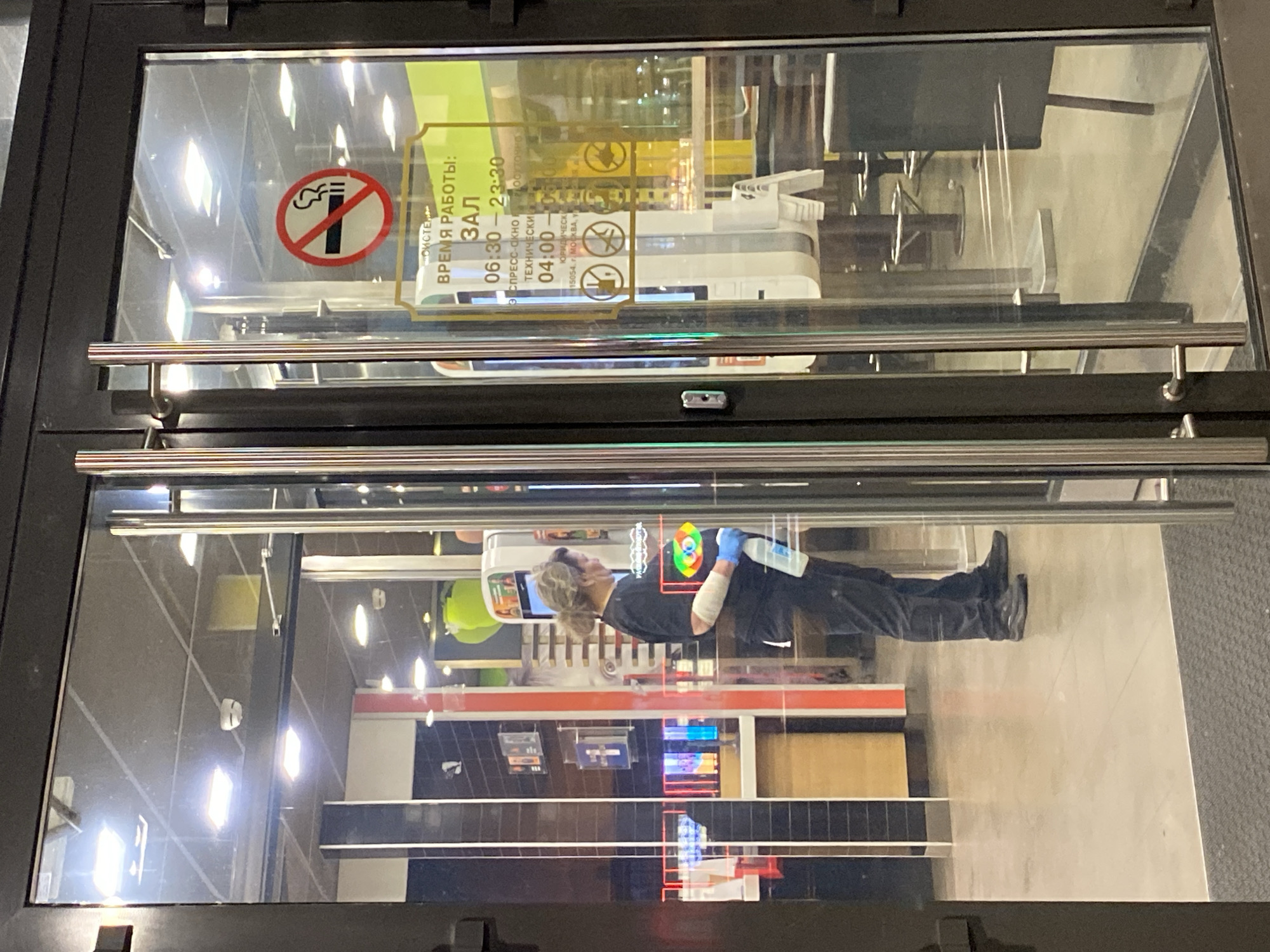The image shows the entrance to a commercial establishment, likely a store, as seen through a set of glass doors framed in dark metal. A "No Smoking" sign and operating hours in Russian are visible on the upper left. The reflection on the glass reveals the interior of the store and a person, presumably an employee or cleaner, wearing dark clothing, blue gloves, and holding a spray bottle. The person appears to be working in the store, based on their attire and actions. While the location cannot be precisely determined from the image, the OCR text visible mentions "Москва" (Moscow), suggesting this is not Saint Petersburg, but rather Moscow, Russia.