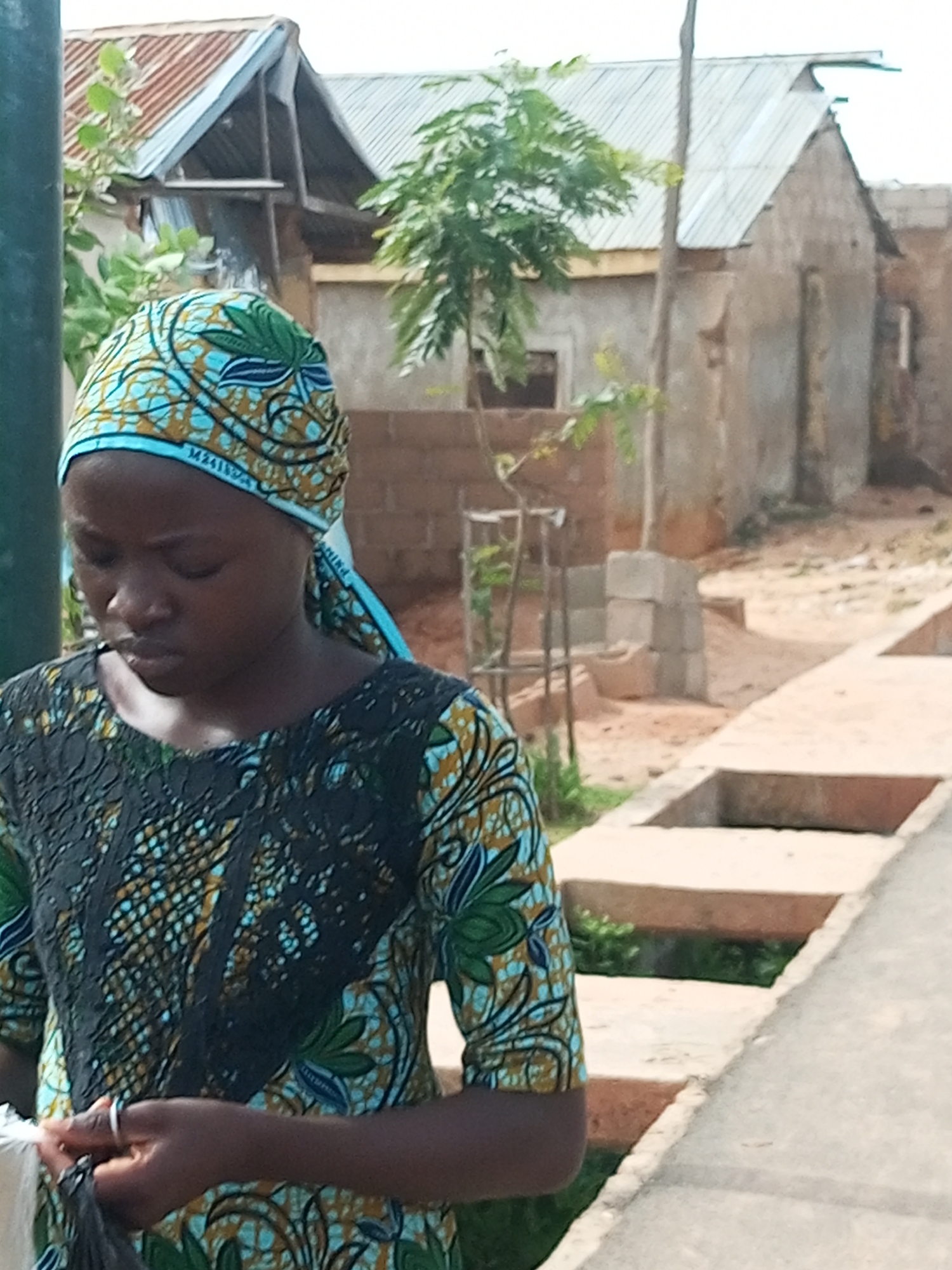 A young woman with dark skin and brown eyes is standing outside, looking down. She is wearing a blue and green headscarf with a geometric pattern and a black and teal patterned dress. Her hands are clasped in front of her, and she is holding a white object. The background shows a dirt path, some buildings with corrugated metal roofs, and a tree. The scene appears to be in a rural or semi-urban area, possibly in Tempure, Nigeria.