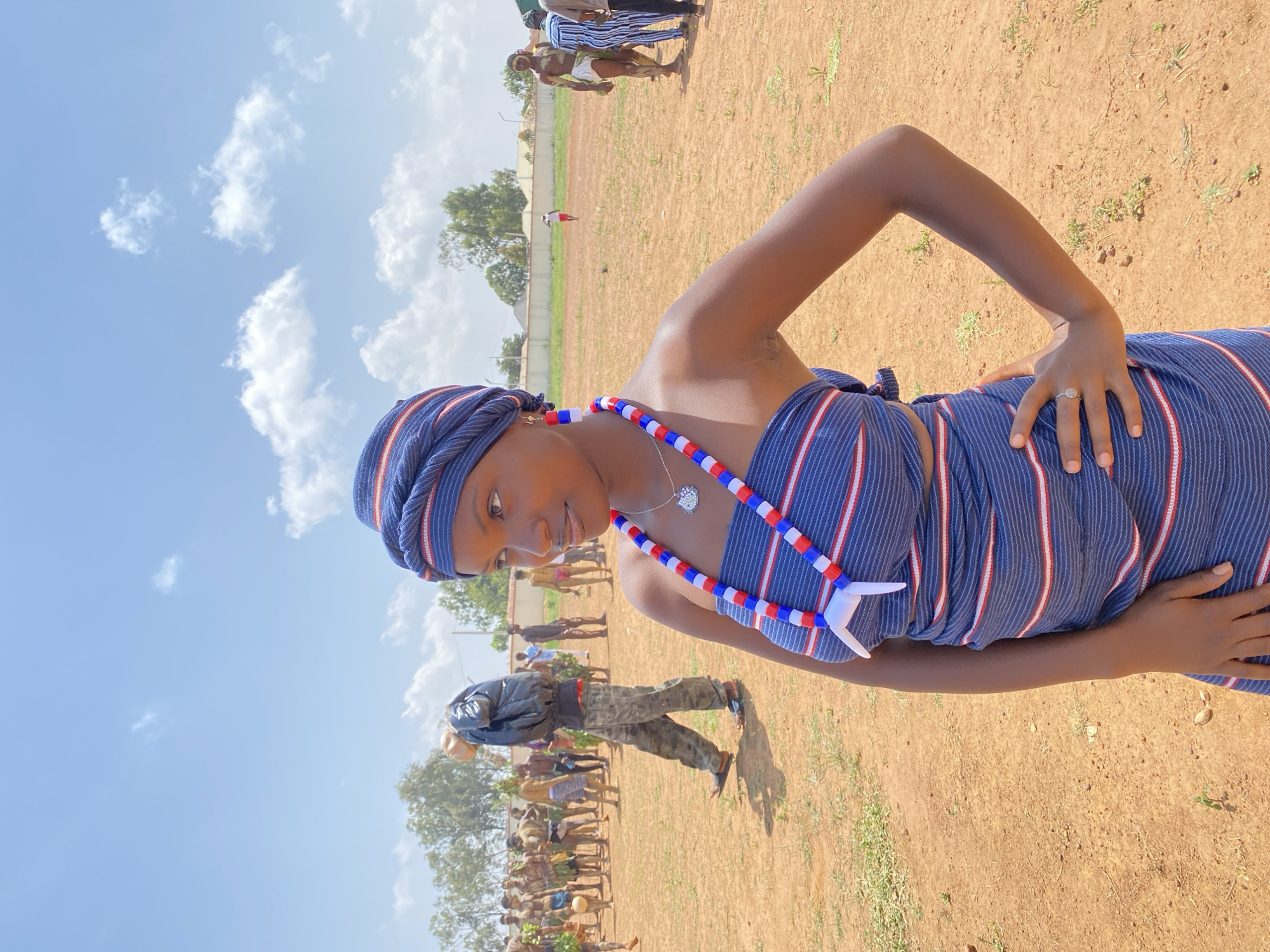 A young woman is the primary subject of the image, standing in an outdoor setting. She is wearing a traditional blue and white striped cloth wrapped around her body and a matching head wrap. A necklace with blue, red, and white beads adorns her neck. She is holding her body in a relaxed pose, with her hand at her waist.
The background reveals a field with sparse grass and trees in the distance. A number of people are gathered in the background, including some wearing similar traditional attire. Based on the attire and scenery, it appears to be a cultural event or gathering in Durimi, Nigeria. The environment is filled with a bright, sunny sky with scattered clouds.
