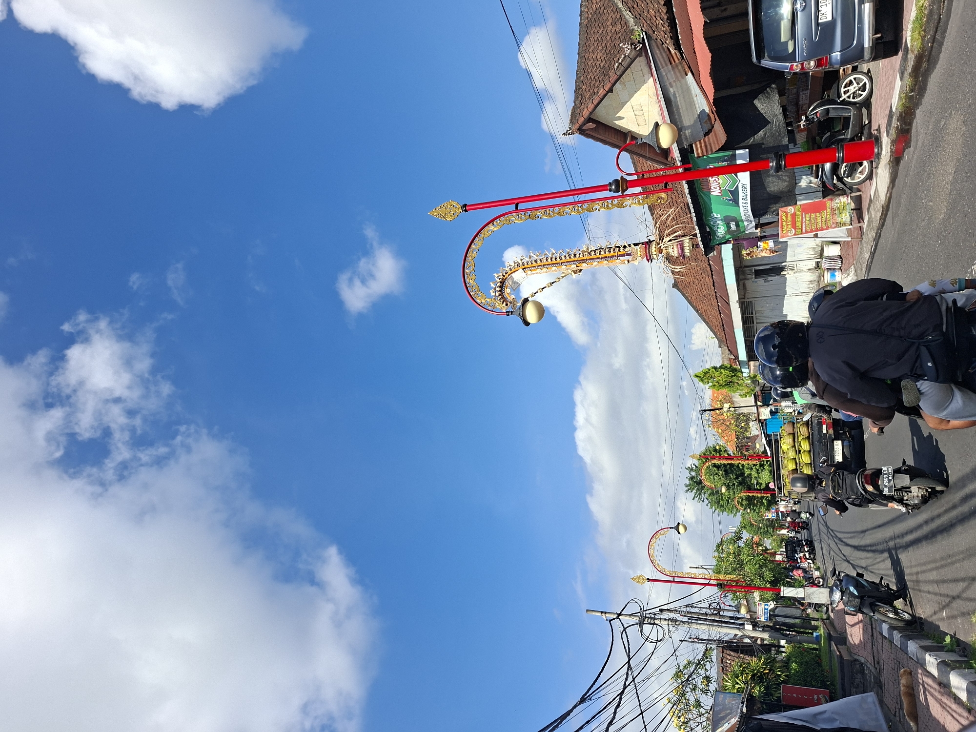 The image shows a street scene in Gianyar, Indonesia, on a bright, sunny day with a blue sky dotted with white clouds. The street is paved and has a slight incline. On the right side of the street, there are buildings with traditional tiled roofs and awnings. One building has a green banner that reads "Nonna's Cake & Bakery". There are several motorcycles and a truck on the road. The truck is carrying what appears to be bundles of yellow produce. In the foreground, two people are riding on a motorcycle, both wearing helmets. On the left side of the street, there is a sidewalk with a few buildings and some greenery. A ginger cat is lying on the sidewalk. Decorative lampposts with red poles and ornate gold accents are visible along the street. The scene depicts a typical day in a busy urban area with local commerce and traffic.