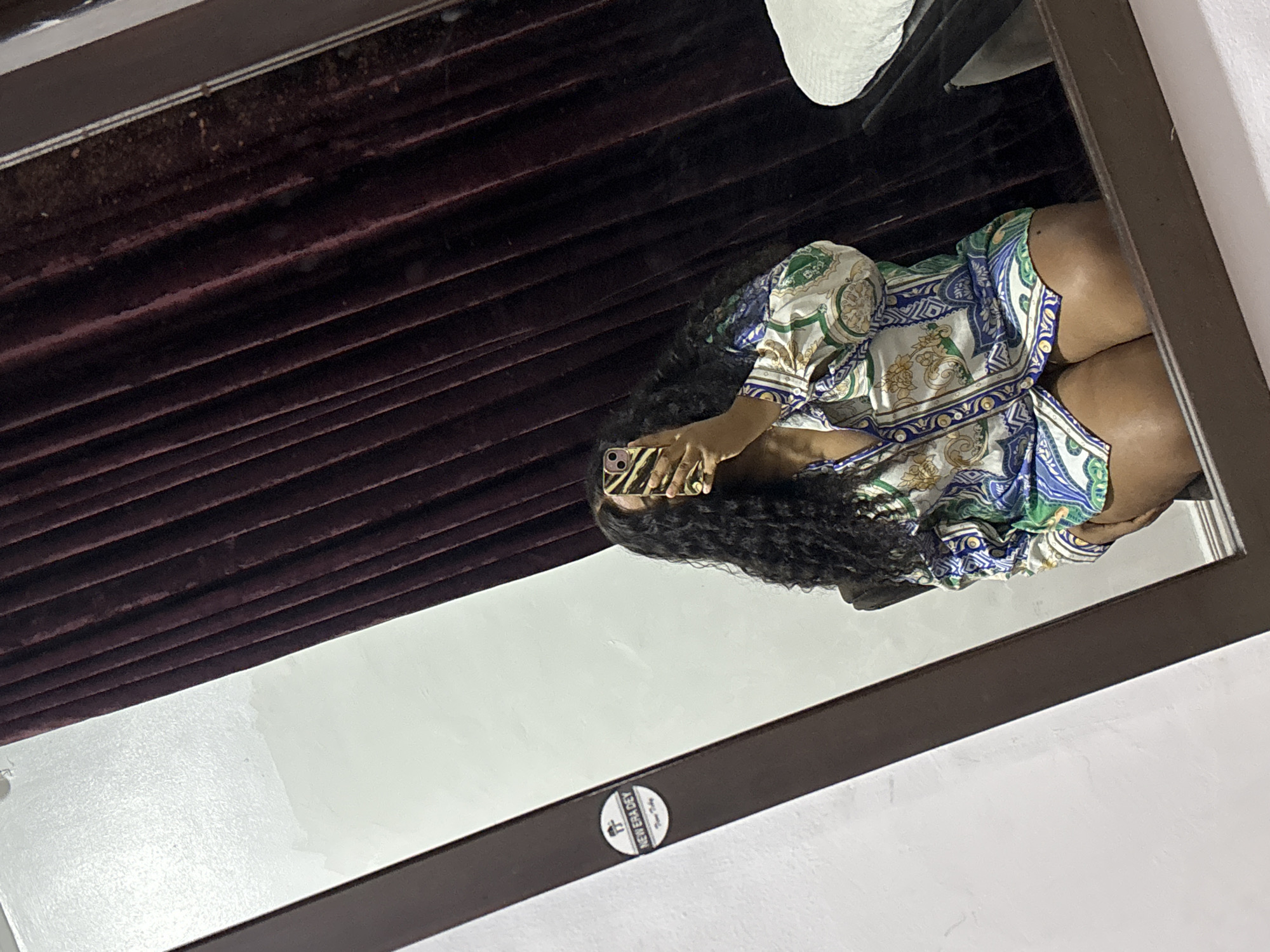 A mirror reflects a person with dark curly hair, wearing a patterned blouse, holding a phone with a zebra-striped case. The reflection also includes a portion of what appears to be a dark-colored Venetian blind and the edge of a white object. The mirror itself has a dark frame. A small round sticker, possibly a brand logo, is visible on the mirror's surface in the lower center. The location is in a room in Port Harcourt, Nigeria.