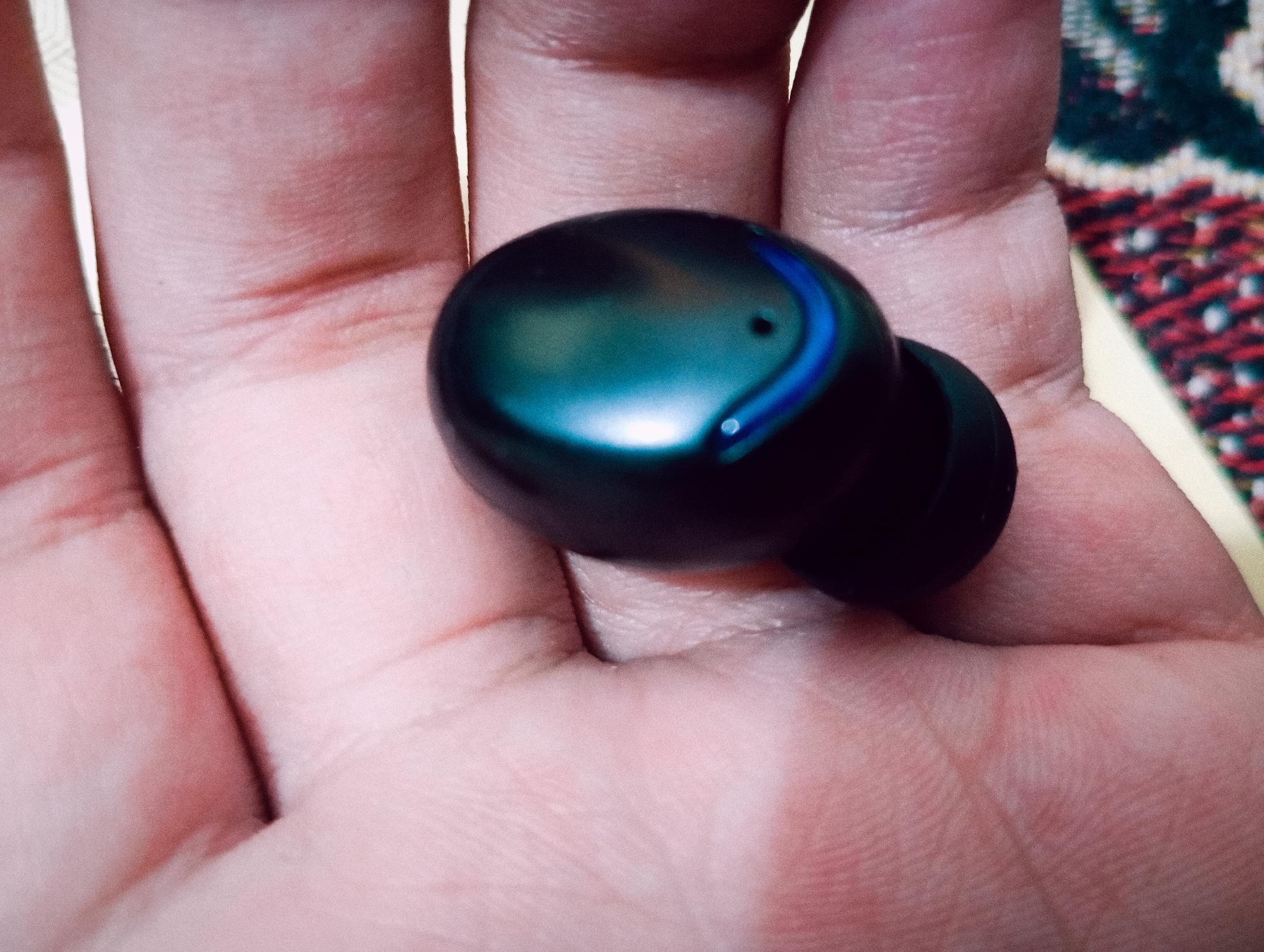 A close-up shot shows a person's hand holding a single black wireless earbud with a blue accent. The earbud is placed in the palm of the hand, which is slightly cupped. The skin of the hand is visible, with some creases and faint red marks. In the background, a blurred colorful patterned fabric is visible. The lighting suggests an indoor setting. The image does not contain information about a city or its location.