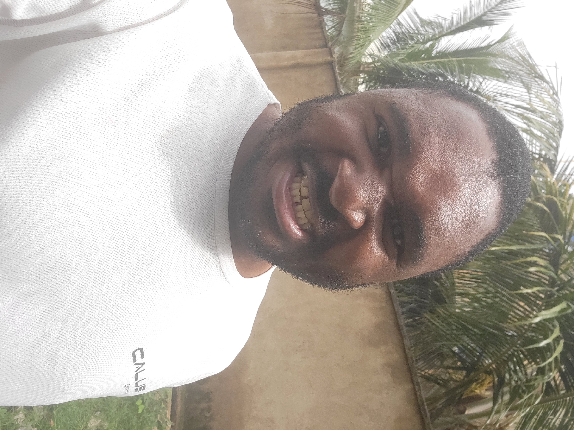 The image is a close-up selfie of a smiling man with dark skin, a beard, and short black hair. He is wearing a white t-shirt with a logo on the lower left. The background consists of a beige wall and palm tree foliage. Given the prompt includes Suleja, Nigeria, it is likely the image was taken there. The lighting is bright, indicating it's likely daytime. The subject's focus is directly at the camera.