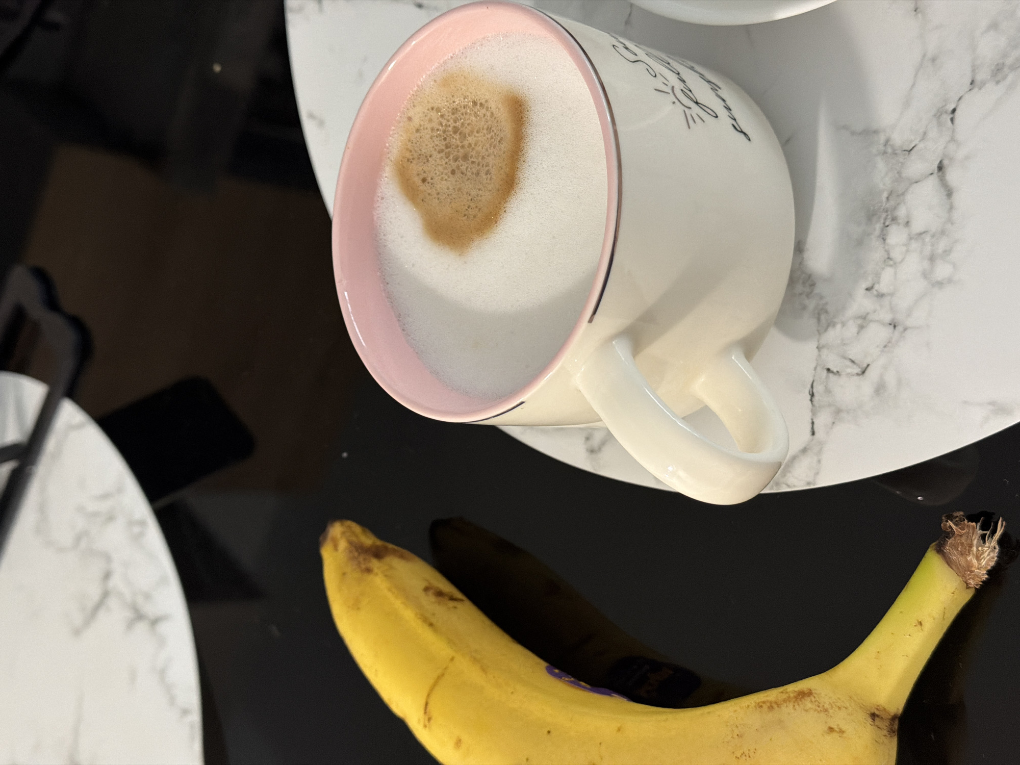 A close-up, eye-level shot shows a ripe yellow banana positioned on the left side of a black glossy surface. The banana has some brown spots and a purple sticker is partially visible on its peel. To the right of the banana, a white ceramic mug with a pink rim rests on a white marble coaster. The mug contains a latte with a frothy layer of milk and a small, off-center patch of darker foam. The mug is decorated with subtle black lettering that appears to say "Sunshine". The background is out of focus, suggesting a shallow depth of field, but hints at a dark reflective surface and portions of white marble placemats or tabletops. The scene suggests a breakfast or snack setting, possibly in a home or cafe environment. There is no indication of Finestrat, Spain.
