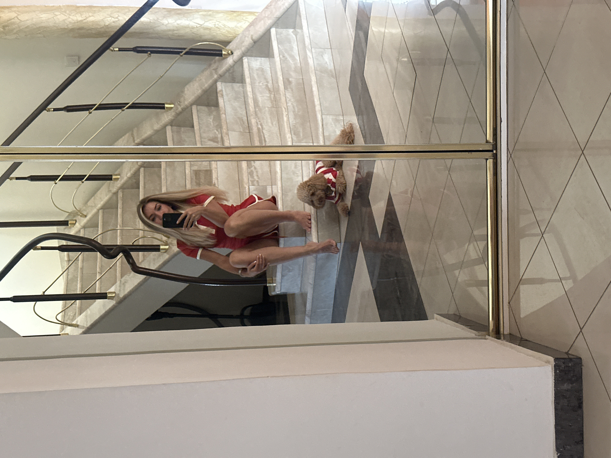 A blonde woman in a red and white dress is sitting on a marble staircase, holding a smartphone to take a selfie. A small dog wearing a red and white striped shirt lies on the floor in front of her. The scene is reflected in a large mirror. The image appears to be taken indoors, possibly in a residential building. The woman's pose suggests she is casual and relaxed. The dog is alert and looking towards the camera. The staircase has dark wooden railings with gold accents. The floor is tiled in a black and white diamond pattern. The environment is clean and well-maintained. There is no visible indication that this scene is located in Armavir, Russia.