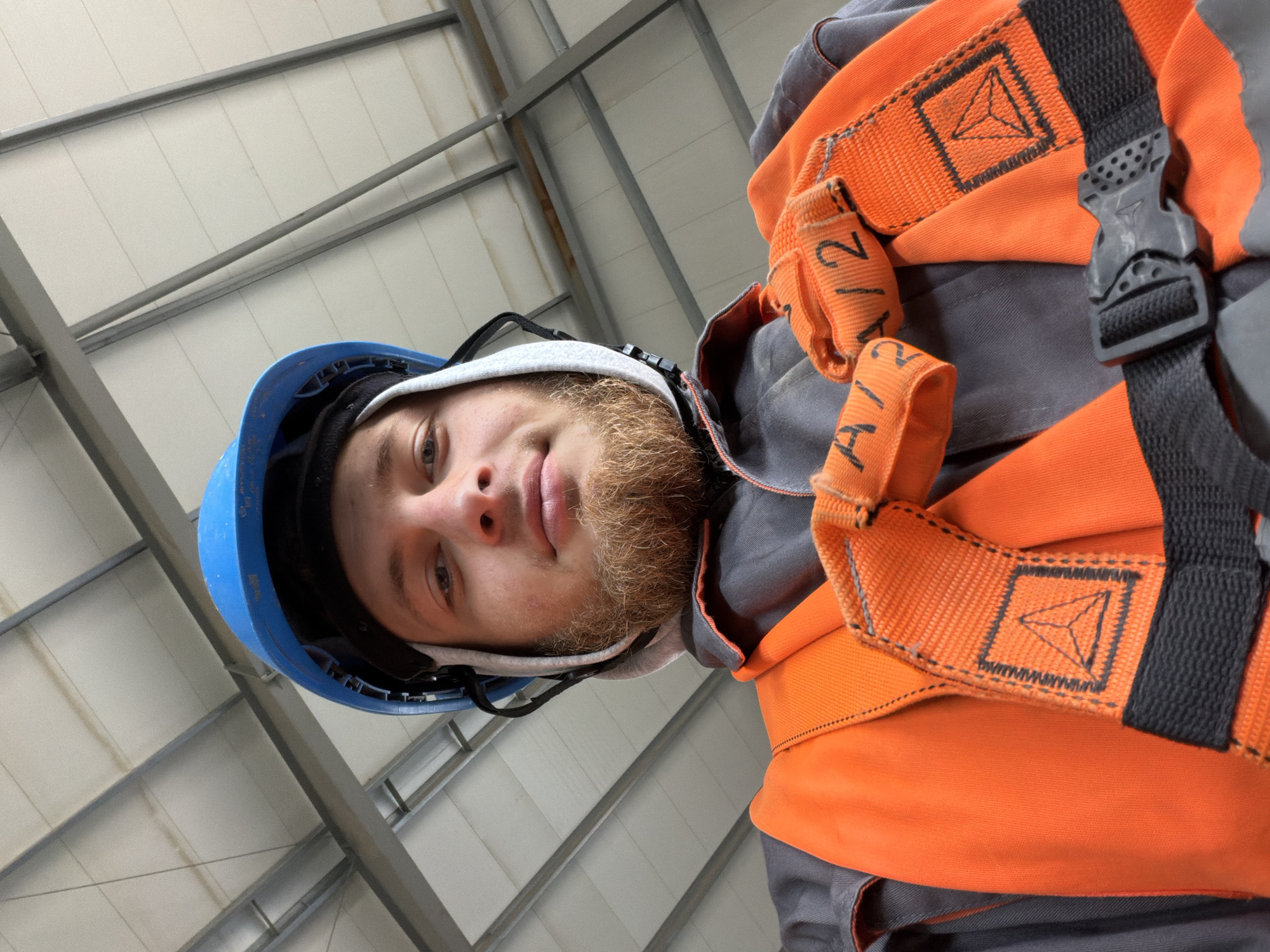 An angled selfie taken from a low perspective shows a young man wearing a blue hard hat, a grey uniform, and an orange safety harness. He has a light-skinned complexion, light blue eyes, and a ginger beard. His hair is also light brown. The background of the image is the interior of a building with a white, insulated ceiling supported by grey metal beams. The overall lighting suggests an indoor, possibly industrial or construction environment. The scene is devoid of any visible city or urban indicators that would allow for specific localization to Janowice, Poland.