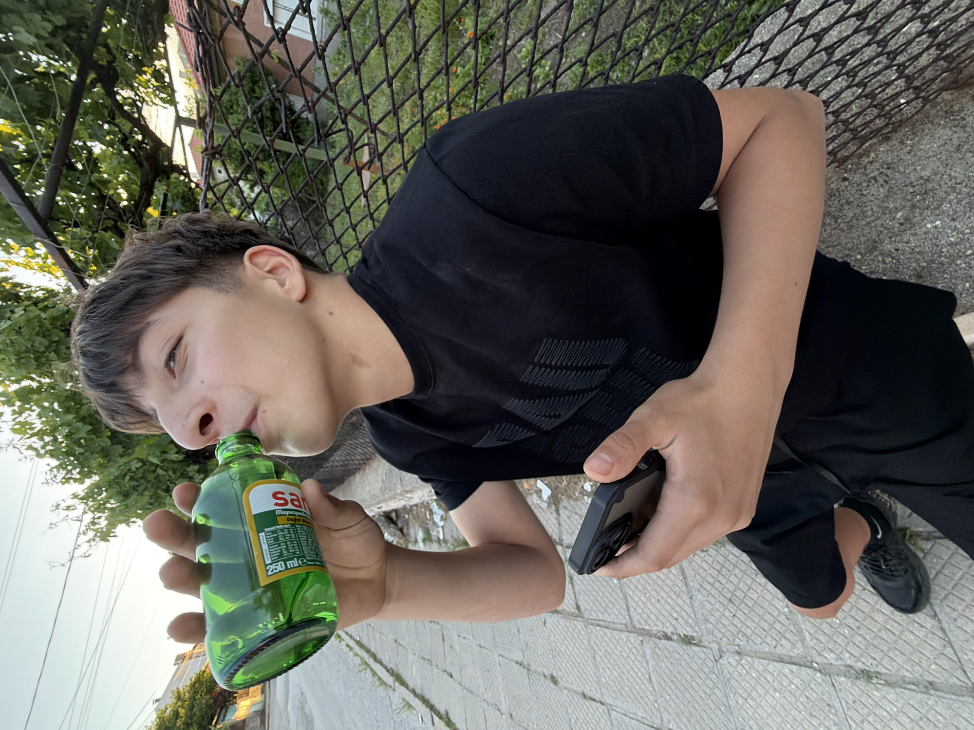 A young male is photographed outdoors in Asenovgrad, Bulgaria. He is drinking from a small, green glass bottle labeled "Salina," which indicates it contains mineral water. The bottle is 250 ml in volume. The male also holds a black cell phone. He is dressed in black athletic clothing. The background includes a chain-link fence, greenery, a building, and a paved sidewalk with grid patterns.