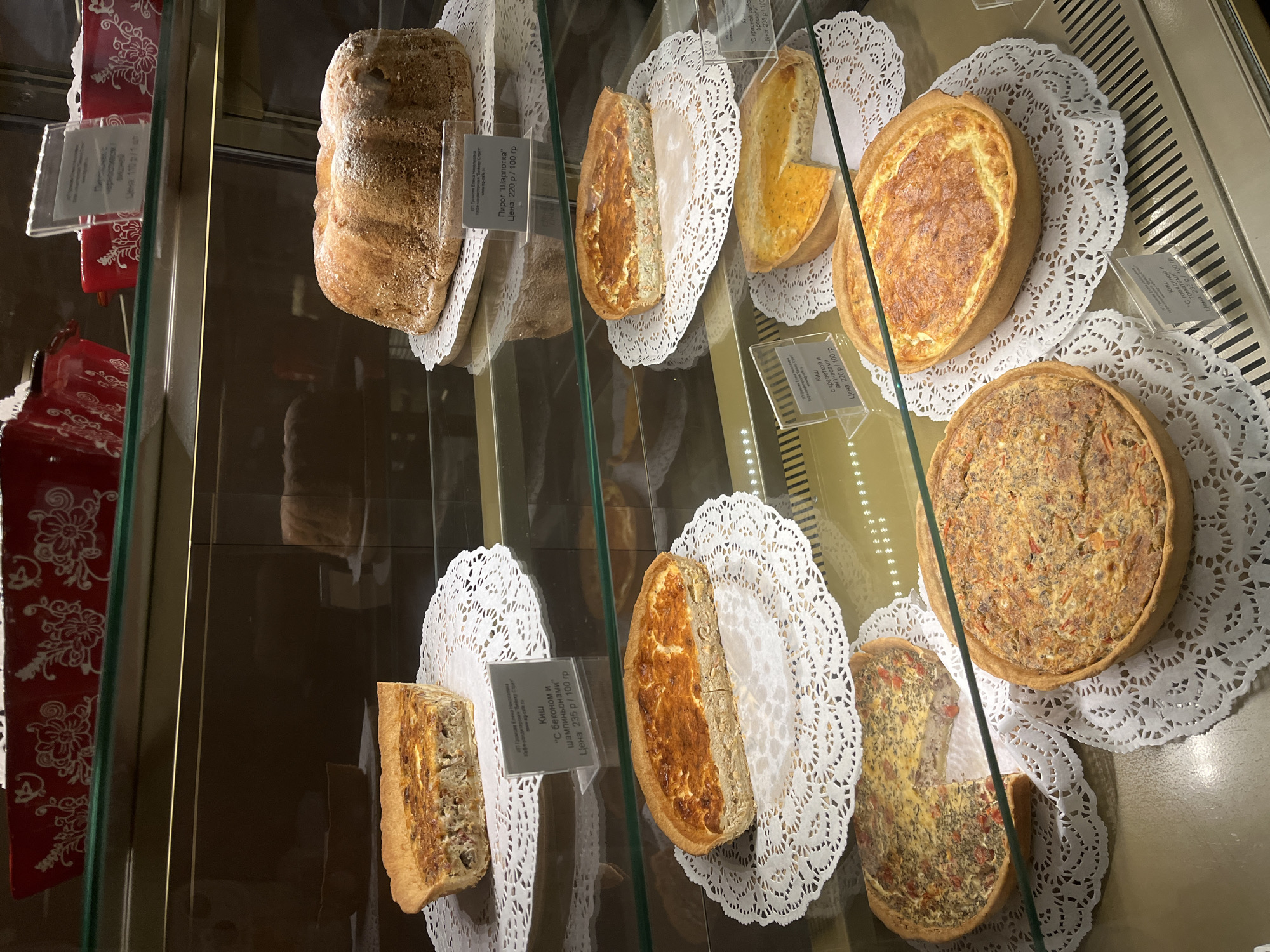 The image shows a display case in a bakery, likely in Petropavlovsk-Kamchatsky, Russia. Various quiches and a bundt cake are arranged on white doilies. Signs in Russian indicate the names and prices of the items. The display case is well-lit, showcasing the baked goods in detail. There are no people visible in the image.