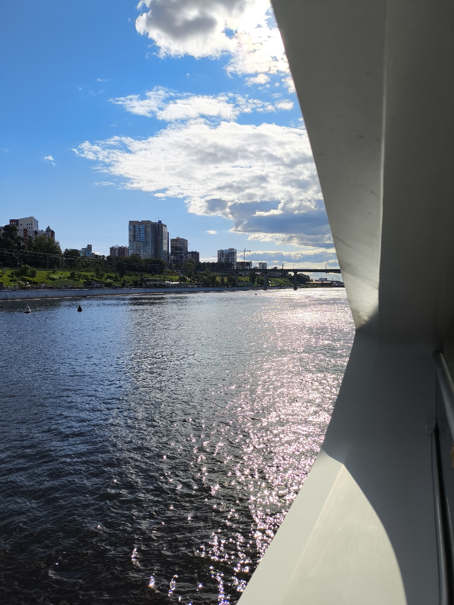 The image captures a view of the Kama River in Perm, Russia, on a bright, sunny day with scattered clouds. The foreground is dominated by a large, light-colored structure, likely part of a building or bridge, which bisects the frame diagonally from the lower right corner upwards. The surface of the Kama River occupies the left and center of the image, reflecting the sunlight with a shimmering, sparkling effect. The water appears calm with gentle ripples.
In the mid-ground, the riverbank is visible, featuring a lush green park area with trees and grass. Beyond the park, a cityscape rises, characterized by several modern high-rise buildings of varying heights and architectural styles. Some buildings are clad in glass, reflecting the blue sky. Construction cranes are visible in the distance, suggesting ongoing development. A bridge spans the river in the far distance, connecting the two sides of the city. Two buoys are positioned in the river, one to the left and another slightly more towards the center. The sky above is a vibrant blue, interspersed with fluffy white and grey clouds, with the sun shining through, creating a bright and airy atmosphere. No people are clearly discernible in the image.