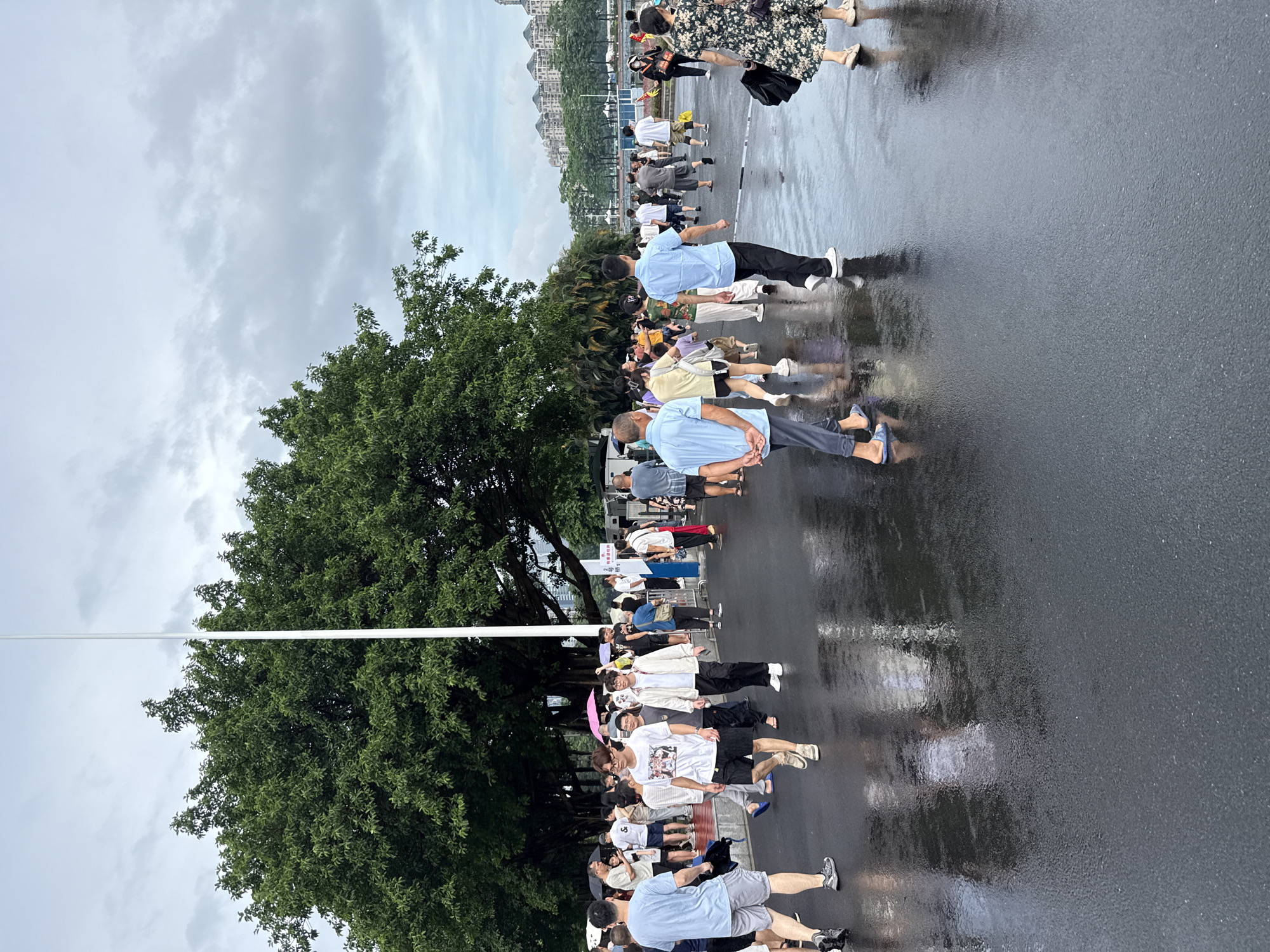 A group of people are walking on a wet pavement in what appears to be a public outdoor area. The ground is reflective, indicating recent rainfall. Several individuals are seen from various angles, some walking towards the camera, others away. They are dressed in casual clothing, including t-shirts, shorts, and pants. A few people are holding umbrellas, suggesting it may have recently rained or is still drizzling. In the background, there are large trees with lush green foliage and a white flagpole. Further in the distance, buildings are visible, and a cloudy sky looms overhead. The overall scene depicts a busy day in an urban environment, possibly Tianhe District, China, with people navigating the conditions after rain.