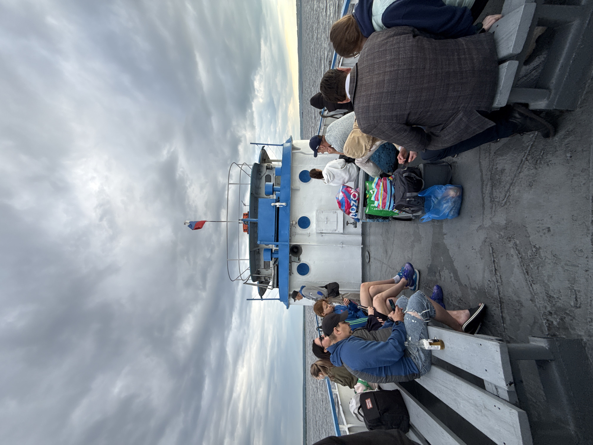 A group of people are seated on benches on the deck of a ferry boat sailing on a body of water under a cloudy sky. The boat has a white superstructure with blue trim and a Russian flag flying from a mast. Several passengers are looking at their phones, while others are looking out at the water or talking to each other. Bags and backpacks are placed on the deck around the passengers. The scene appears to be in the city of Kazan, Russia, judging by the presence of the Russian flag and the typical architecture of such ferries on the Volga River.