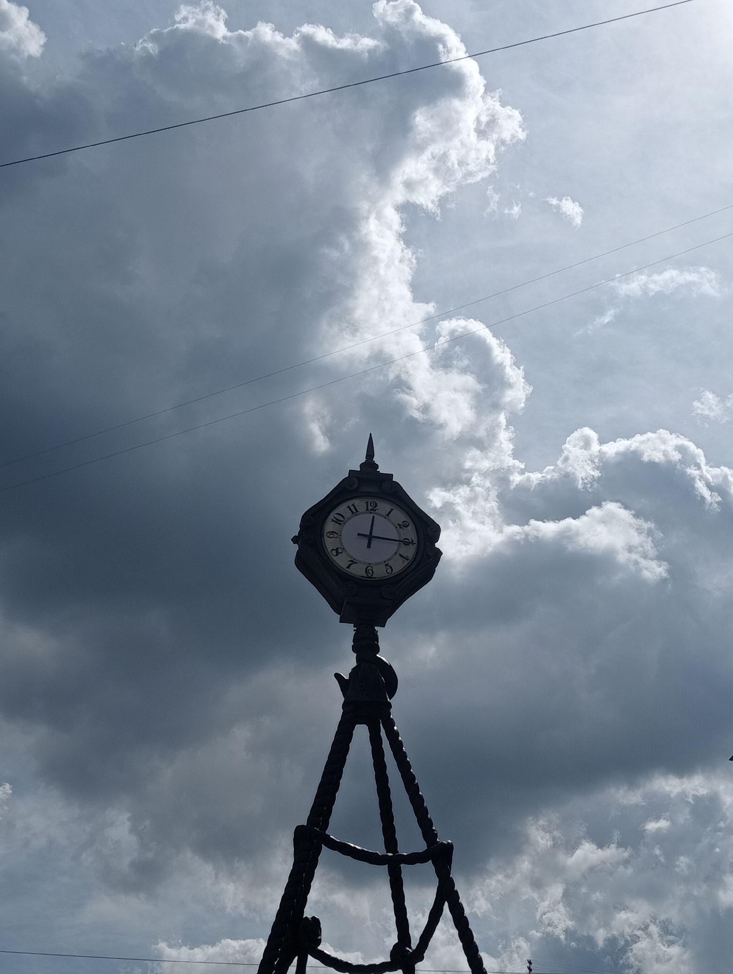 The image captures a silhouette of a clock tower against a dramatic sky filled with dark, ominous clouds and bright, puffy white clouds. Two thin wires are visible stretching across the sky, suggesting an urban setting with overhead power lines. The clock face on the tower clearly displays the time as approximately 11:52. The base of the tower appears to be made of intricate, dark metalwork, resembling twisted ropes or cables. The overall atmosphere is one of impending weather, with the contrast between the dark clouds and the illuminated clock tower creating a striking visual. This scene is reminiscent of the iconic clock towers found in various locations throughout Saint Petersburg, Russia, often adorning public squares or historical buildings.
