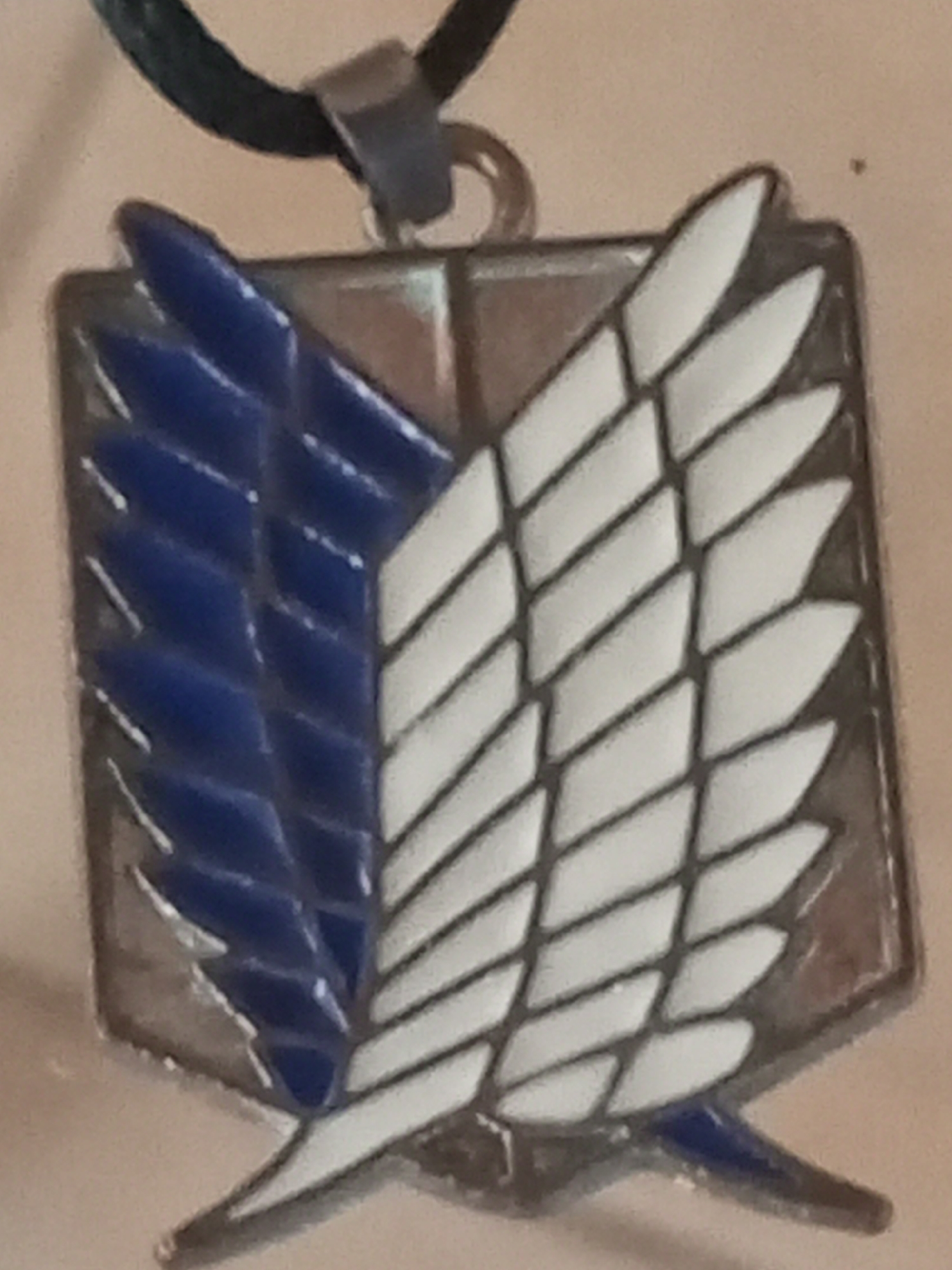 The media file shows a close-up of a pendant, likely a necklace, featuring the emblem of the Survey Corps from the anime series "Attack on Titan." The emblem is shield-shaped and displays a stylized pair of wings. The left wing is colored blue, and the right wing is colored white. The wings are outlined in black, and the shield itself has a dark border. The pendant is suspended from a black cord with a metal clasp. The background is a light, neutral color, possibly skin or a fabric surface, and is out of focus. There is no visible text in the image. The lighting appears to be indoor lighting, with no strong shadows. The overall impression is of a personal item, possibly worn as a piece of fan merchandise. There are no people, specific scenes, or events depicted, only the object itself. The location context of Altagracia, Venezuela, does not provide any visual cues within the image.