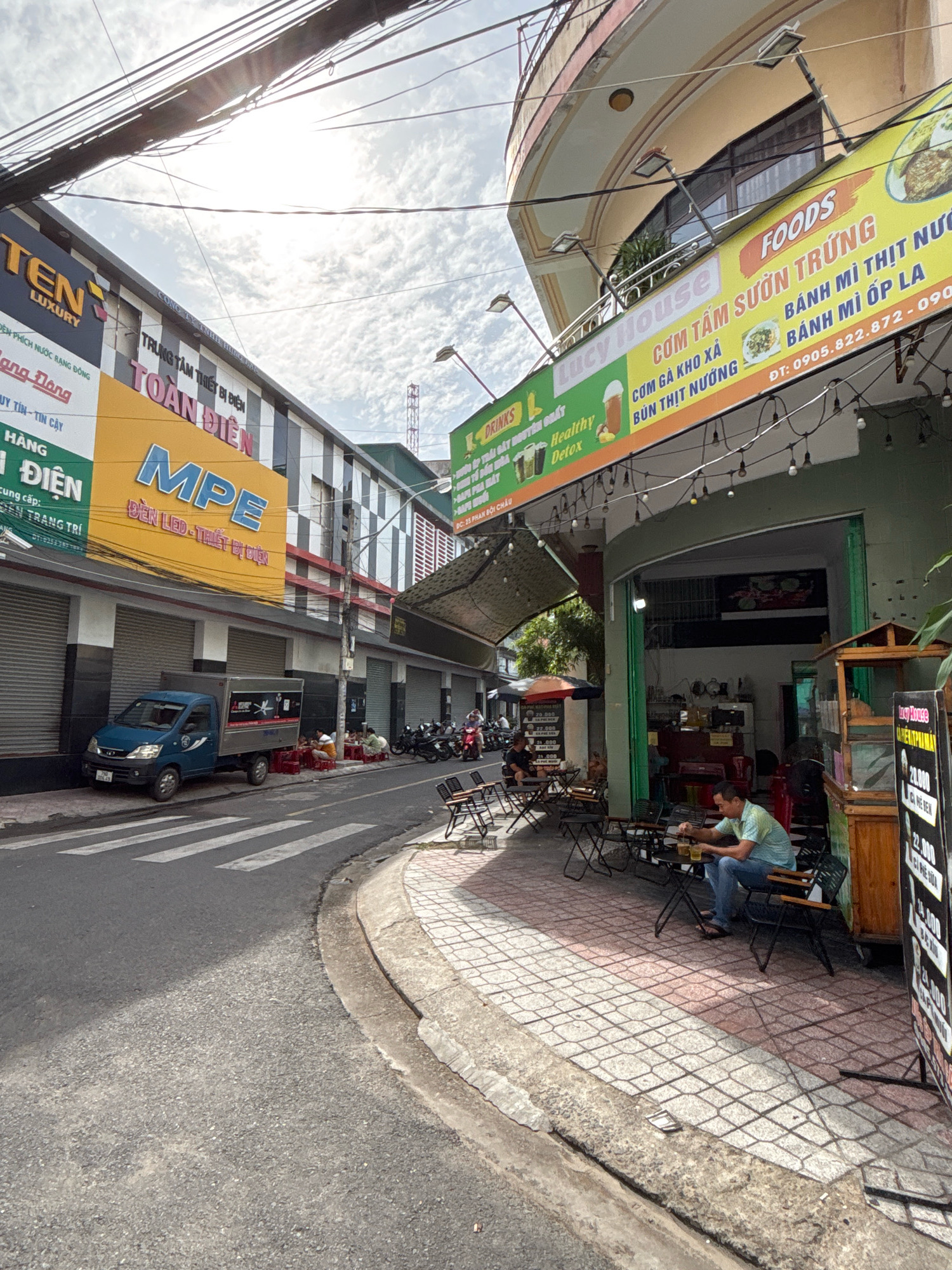 The image depicts a street scene in Nha Trang, Vietnam, on a cloudy day. The foreground shows a crosswalk and a section of the road. On the right side of the street, a man is seated at an outdoor cafe table, holding a glass. Several other empty tables and chairs are arranged outside the establishment, which appears to be a food and drink vendor. To the left of the street, a blue truck is parked near a building with a large yellow and orange sign. Further down the street, other businesses with various signs are visible, including one with the words "FOODS" and "LUCY HOUSE." There are also several motorcycles parked along the sidewalk, and a few people can be seen in the distance. Overhead, a complex network of electrical wires spans across the scene.