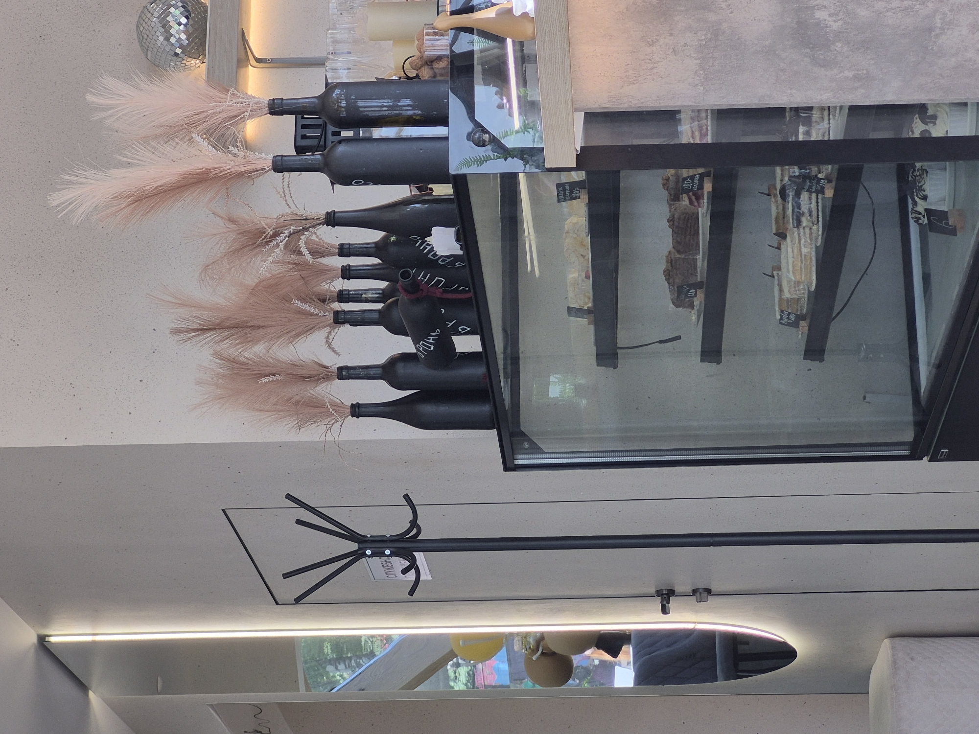 This image captures an indoor scene with a combination of decorative and functional elements. The main focus is a glass display case filled with what appears to be various food items or ingredients. Adorned near the display case are several dark-colored bottles, possibly wine bottles, each decorated with light pink, feathery plumes. A coat rack with multiple arms is also visible, suggesting a public or semi-public space. A reflective surface or mirror below the light fixture hints at people being present, including one person wearing blue. The overall environment suggests a cafe or boutique setting. The city location is Unknown, Unknown.