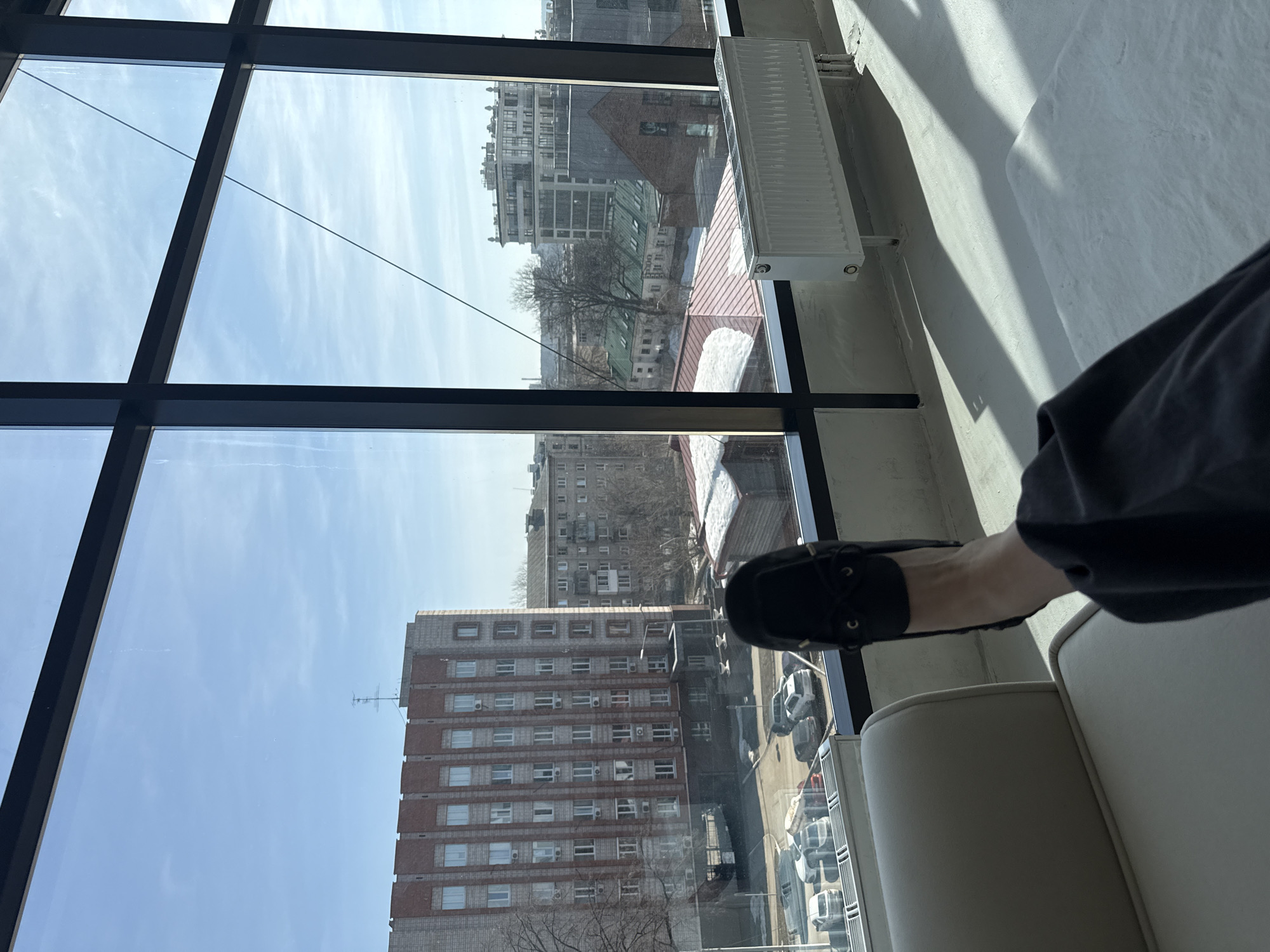A low-angle, indoor shot presents a cityscape viewed through a large, multi-paned window. The window frame is black. Outside, the scene is bathed in daylight, under a sky dotted with wispy clouds. A tall, brick-faced building with numerous windows dominates the lower portion of the view, while other buildings and structures stretch into the distance. A radiator is mounted to the wall next to the window.
In the foreground, a person is seated on a light-colored piece of furniture. The person is wearing dark-colored pants and a black loafer.
Based on visual details such as the architectural style of the buildings and the presence of Russian license plates on the parked cars, the location is identified as Izhevsk, Russia.