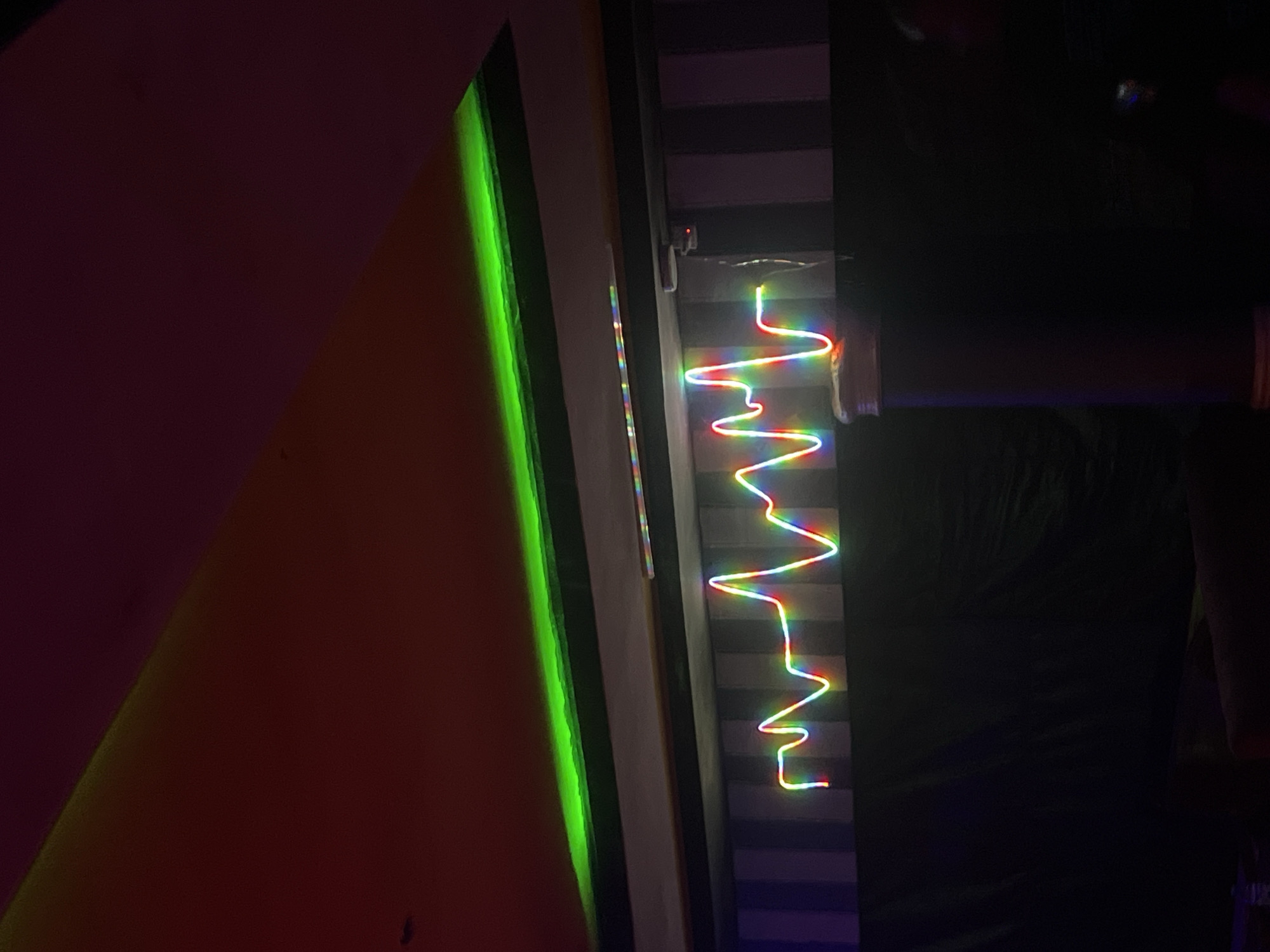 The image shows a room with a ceiling that has a red gradient lighting. A green strip light is embedded along the edge of the ceiling. Below the ceiling, there is a neon light display in the shape of a heartbeat or waveform, illuminated with rainbow colors. This neon sign is mounted against a wall with vertical stripes in alternating light and dark gray. The lower part of the image is dark, showing a black curtain and a dark cylindrical object on the right. There are no people clearly visible in the image. The scene appears to be indoors, possibly a lounge, club, or entertainment venue. The location in Durimi, Nigeria cannot be determined from the image alone.
