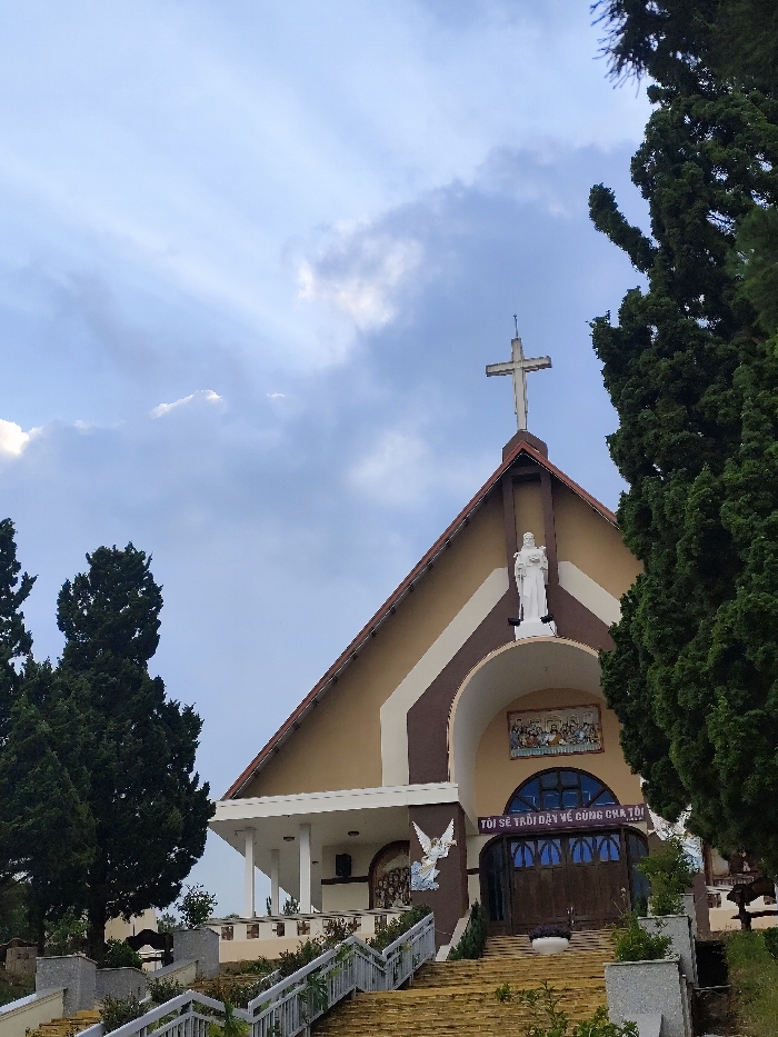 This image depicts the Domaine de Marie Church, also known as Mai Anh Church, located at 01 Ngô Quyền Street, Phường 6, Thành phố Đà Lạt, Lâm Đồng, Vietnam.
The scene presents an upward view of the church's facade under a partially cloudy, light blue sky. The church features a steep, reddish-brown tiled roof culminating in a white cross at its apex. Below the cross, a white statue of a female figure, presumably Mary, stands within a recessed niche. The main facade is primarily painted a light beige/tan with brown and white architectural trim. A large arched entrance dominates the lower central part of the facade. Above the arch, a colorful mosaic panel depicts a religious scene, likely "The Last Supper." Below the mosaic and above the dark wooden double doors, a banner with Vietnamese text reads "TÔI SẼ TRỞI DẬY VỀ CÙNG CHA TÔI." To the left of the main entrance arch, a white relief sculpture of an angel with outstretched wings is affixed to the wall. Further left, a covered porch with white pillars extends from the building.
A wide, multi-tiered staircase, appearing golden-yellow from discolored steps, leads directly up to the church entrance. White railings with grey posts line both sides of the staircase. Green foliage and shrubs are planted alongside the stairs and on adjacent terraced areas. Tall, dark green coniferous trees, resembling cypress or pine, frame the church on both the left and right sides, partially obscuring the upper portions of the sky. No individuals are visible within the frame.