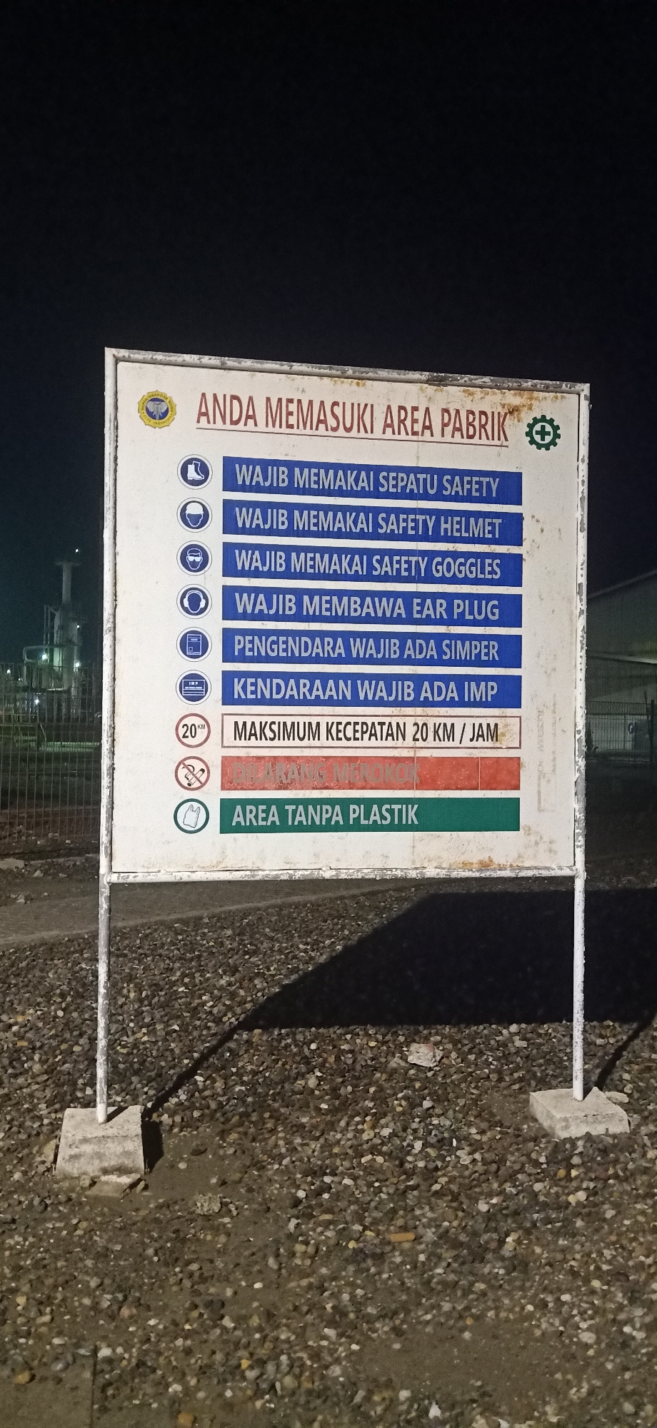 The image shows a large white sign with a blue and red border, situated on two metal poles anchored in concrete bases. The sign is located in Palohlada, Indonesia, at what appears to be an industrial site, evidenced by the presence of factory structures and a chain-link fence in the background. The sign is informative, detailing safety regulations for entering a factory area.
The main text at the top of the sign reads "ANDA MEMASUKI AREA PABRIK" which translates to "YOU ARE ENTERING THE FACTORY AREA." Below this, a list of mandatory safety requirements is presented with corresponding icons:
* "WAJIB MEMAKAI SEPATU SAFETY" (Must Wear Safety Shoes) - Icon of a boot.
* "WAJIB MEMAKAI SAFETY HELMET" (Must Wear Safety Helmet) - Icon of a helmet.
* "WAJIB MEMAKAI SAFETY GOGGLES" (Must Wear Safety Goggles) - Icon of safety goggles.
* "WAJIB MEMBAWA EAR PLUG" (Must Bring Ear Plug) - Icon of headphones.
* "PENGENDARA WAJIB ADA SIMPER" (Riders Must Have SIMPER) - Icon of a document with a checkmark.
* "KENDARAAN WAJIB ADA IMP" (Vehicles Must Have IMP) - Icon of a document labeled "IMP".
* "MAKSIMUM KECEPATAN 20 KM / JAM" (Maximum Speed 20 KM / Hour) - Icon of a speed limit sign with "20 KM".
* A red section with crossed-out cigarette icon indicates "DILARANG MEROKOK" (No Smoking).
* "AREA TANPA PLASTIK" (Plastic-Free Area) - Icon of a plastic bag with a line through it.
The sign is illuminated, likely by artificial light, and the surrounding ground is covered in gravel and some puddles, suggesting recent rain or damp conditions. The overall scene conveys a message of strict safety protocols within the industrial environment.