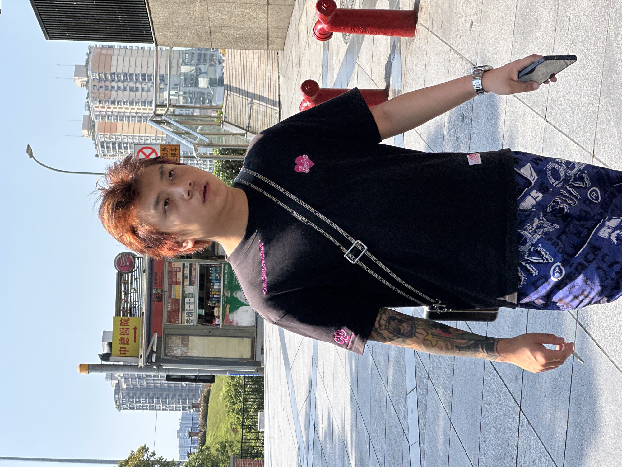 An image depicts a person standing outdoors, likely in an urban setting based on the presence of buildings and infrastructure in the background. The individual has reddish-brown hair and fair skin. The person is wearing a black T-shirt with designs on it and dark blue patterned shorts. They are holding a cell phone in one hand and a cigarette in the other. A shoulder bag is slung across their body.
The background includes a bus stop with signage and a red fire hydrant. Buildings of varying heights can be seen in the distance, as well as a glimpse of greenery. The lighting suggests it is daytime. The OCR text on the bus stop signage reads "中德醫院" (Zhongde Hospital), hinting at a location in China. Based on the signage around the bus stop and the building architectural features, the image is likely taken in the city of Yuzhong District, China.