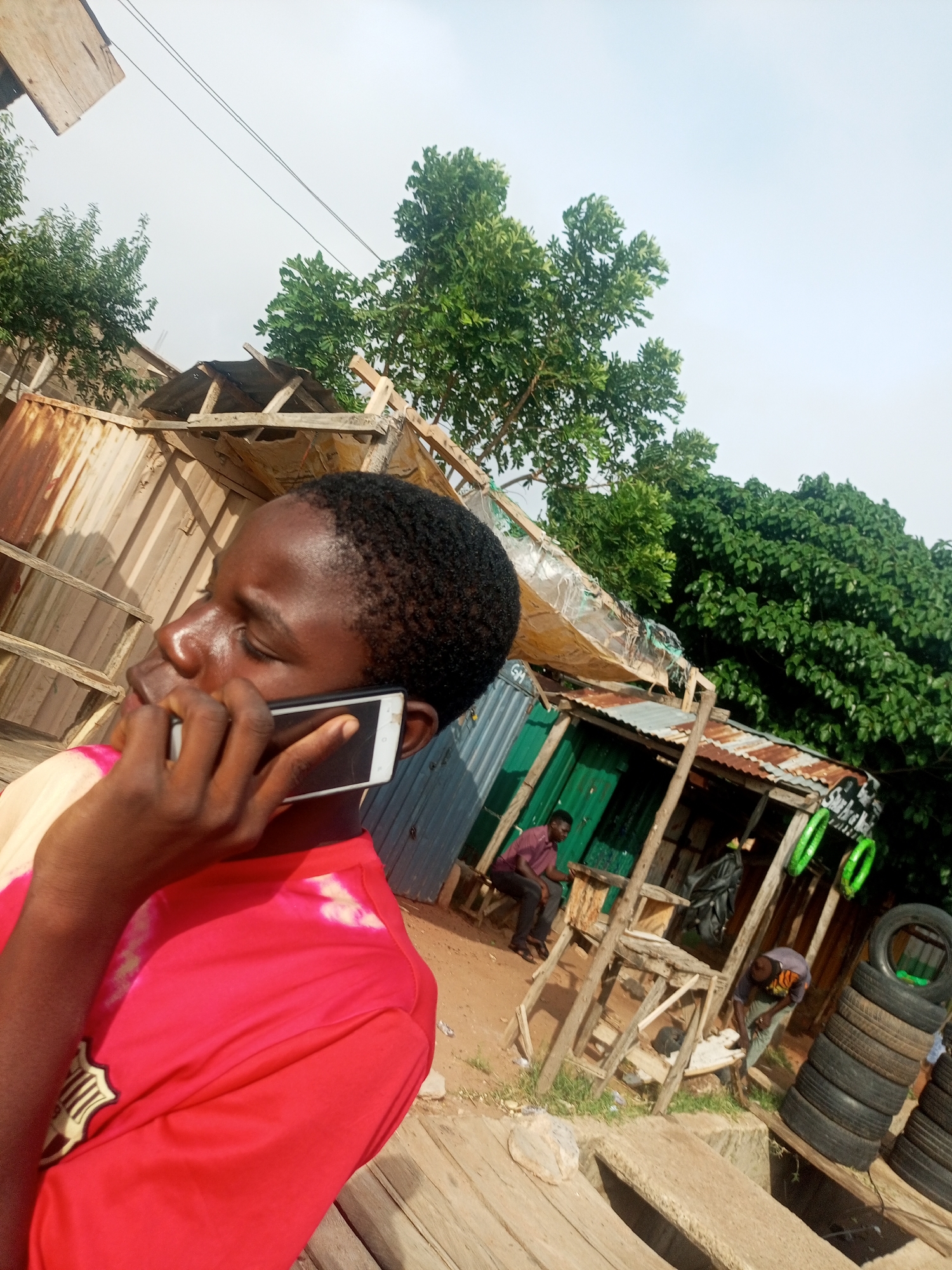 A young Black person in a red shirt is on a phone call, holding a white smartphone to their ear. The person is looking to the left, with their eyes closed, suggesting concentration or perhaps emotion. The background depicts a scene that appears to be a makeshift market or workshop in Tumfure, Nigeria. There are structures built from corrugated iron and wood, with some roofing made of tarpaulin. Several people are visible in the background: one man in a maroon shirt is sitting on a bench, another person in a grey shirt with an orange and black pattern is bent over attending to something, and a third person in a grey and white shirt is also visible. A stack of tires is prominently displayed on the right side of the image. Lush green trees are seen in the background, indicating an outdoor setting. The sky is overcast, suggesting it might be a cloudy day. The overall atmosphere appears casual and industrious. There is some illegible text on a sign in the background.