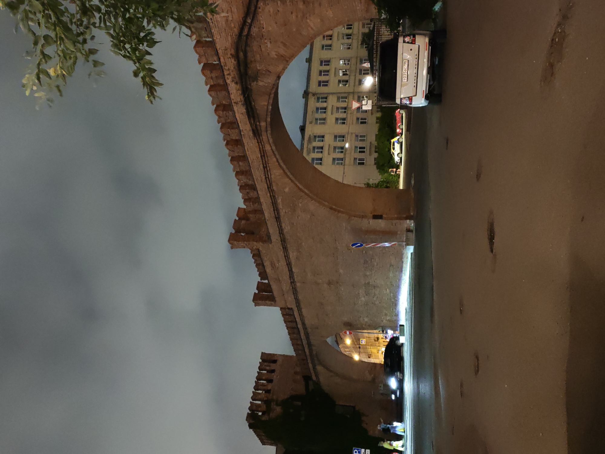 The image depicts a night scene in Saint Petersburg, Russia, featuring an ancient brick archway and wall, likely part of a historical fortification or structure. The architecture is characterized by crenellations along the top of the wall. Beneath the arch, a street is visible with vehicles and a multi-story building in the background.
In the foreground, a white Range Rover is parked on the right side of the frame, with its license plate visible. The street surface shows signs of wear and tear, with several potholes. To the left, under the archway, a black car is driving away, its headlights illuminating the road. Two individuals, one in a yellow garment and another in blue, are standing on the sidewalk to the left. Further down the street, a white car with a blue and yellow stripe on its side and a red sports car are parked.
The sky is overcast and appears dark, suggesting it is nighttime. A tree with green foliage is visible in the upper right corner of the image, partially obscuring the sky. The overall atmosphere is illuminated by artificial light sources, including streetlights and the headlights of vehicles. The location is identifiable as Saint Petersburg due to the architecture and the presence of a license plate format common in Russia.